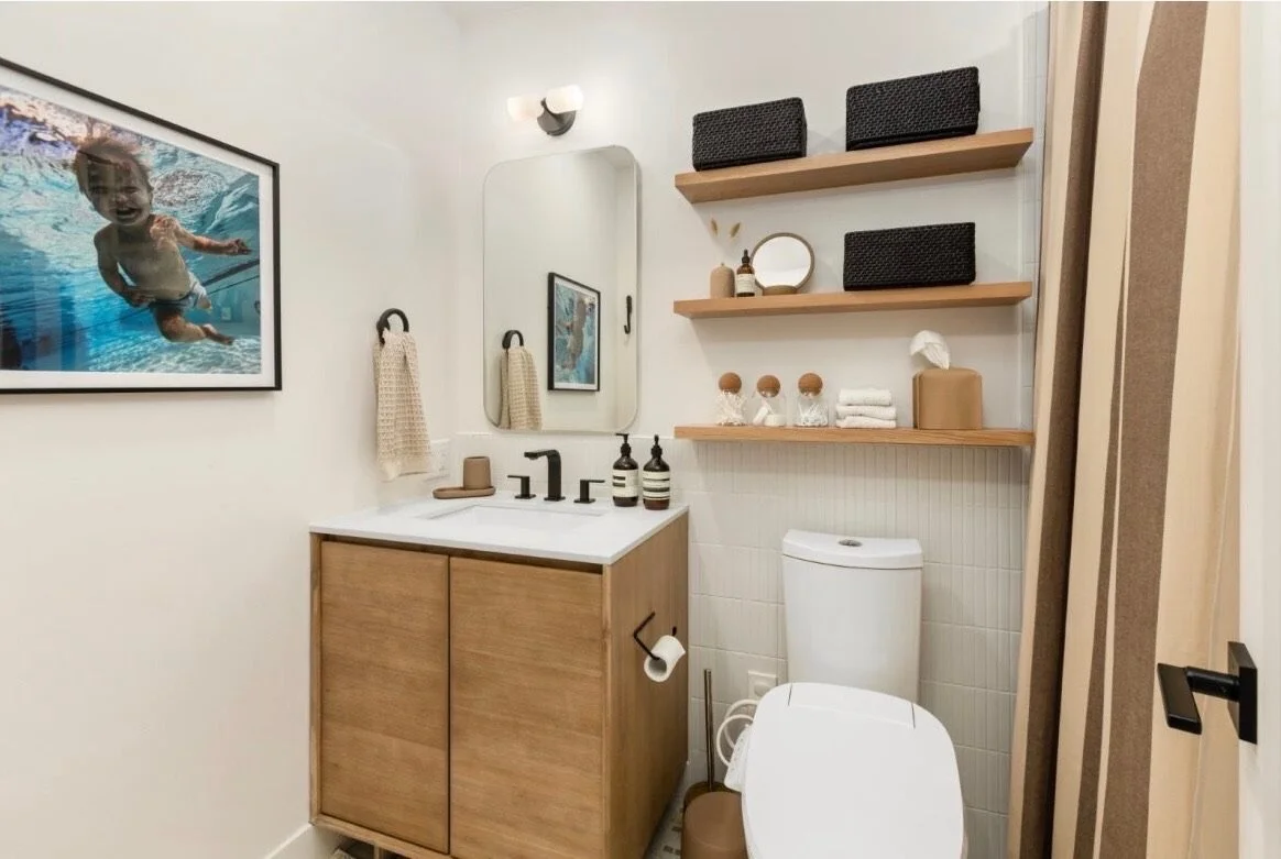 A modern bathroom with a black faucet, wooden vanity with a white countertop, a large mirror, wall-mounted light, towels, decorative items, floating wooden shelves with storage baskets, framed photo of a child swimming, and a white toilet.