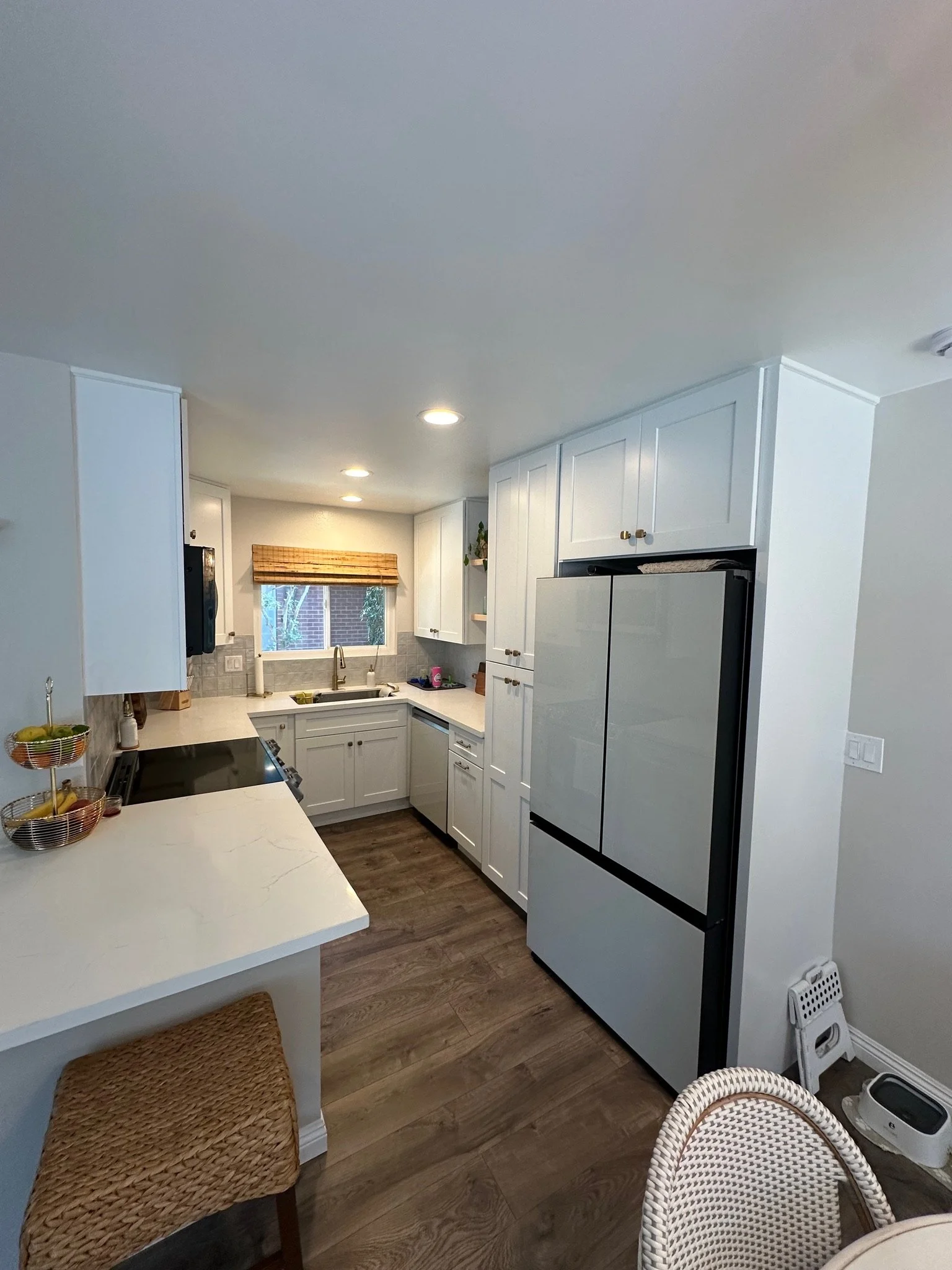Kitchen with white cabinets, a gray refrigerator, and wood flooring, featuring a window with a bamboo blind above the sink.
