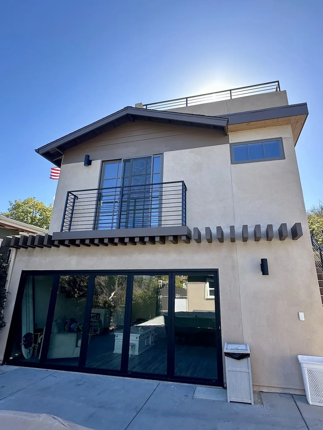 A modern multi-story house with a large glass sliding door on the ground floor, a small balcony on the second floor with black metal railings, and an outdoor staircase on the right side. The house has a beige exterior with dark accents and a sloped roof.