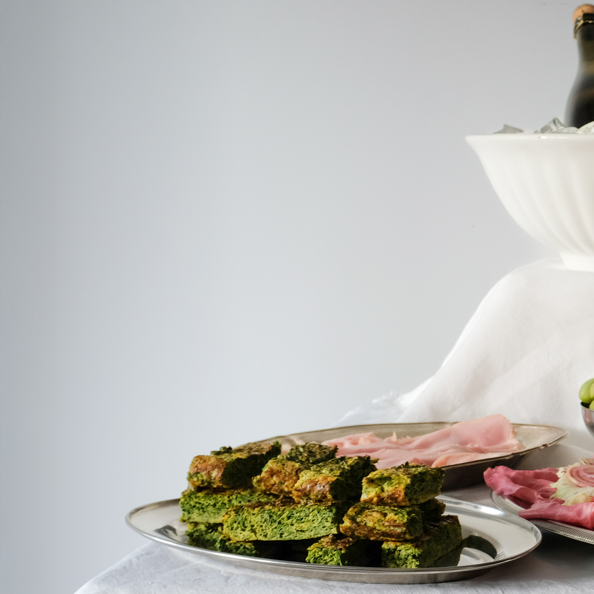 A silver platter with green spinach and meat lasagna pieces, and a silver tray with sliced ham in a white bowl near blurred other food items on a table with a white tablecloth.