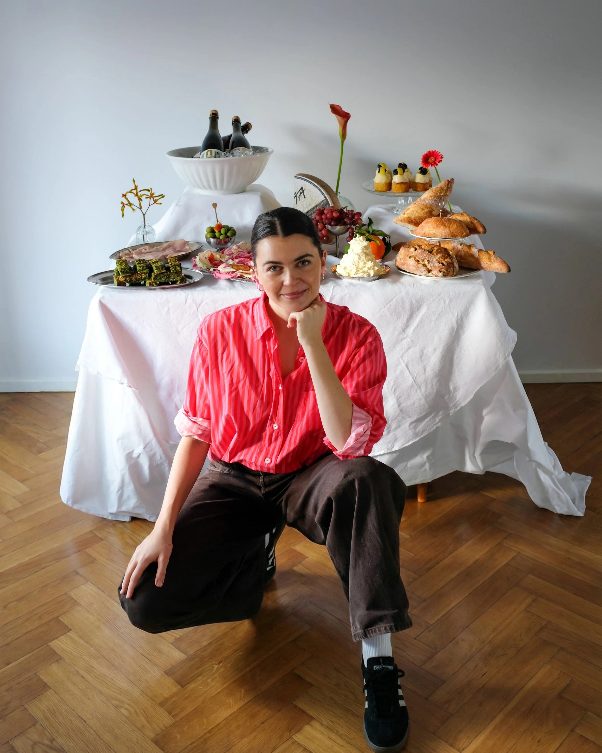 A woman, Larissa Sterchi, in a red and white striped shirt kneels on the floor in front of a table with assorted food and drinks, including bottles, desserts, bread, fruits, and cheese, in a room with wooden flooring.