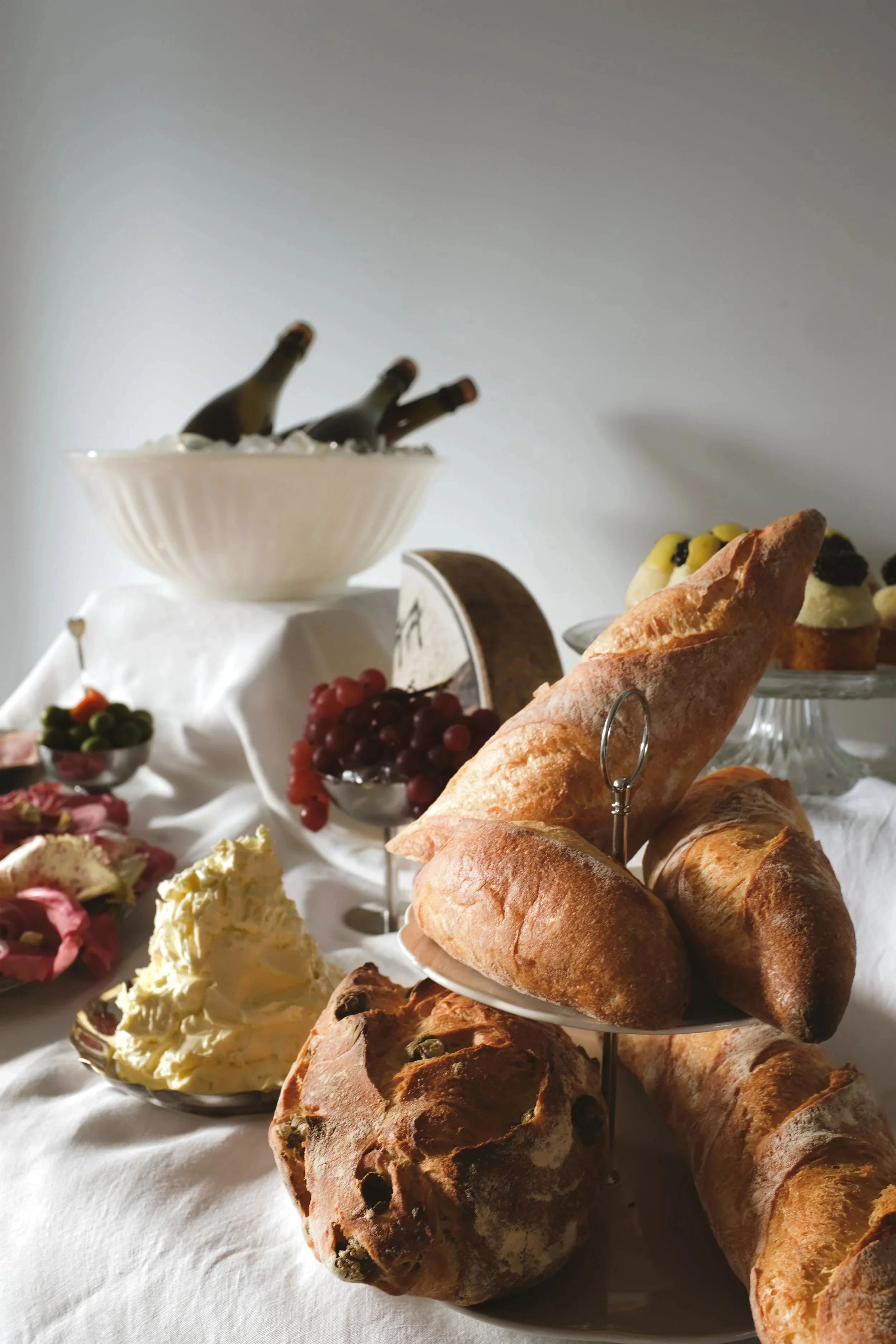 A variety of brunch items including freshly baked baguettes and pastries on a tiered tray, bowl of whipped butter, grapes, and other dishes, set on a white tablecloth.