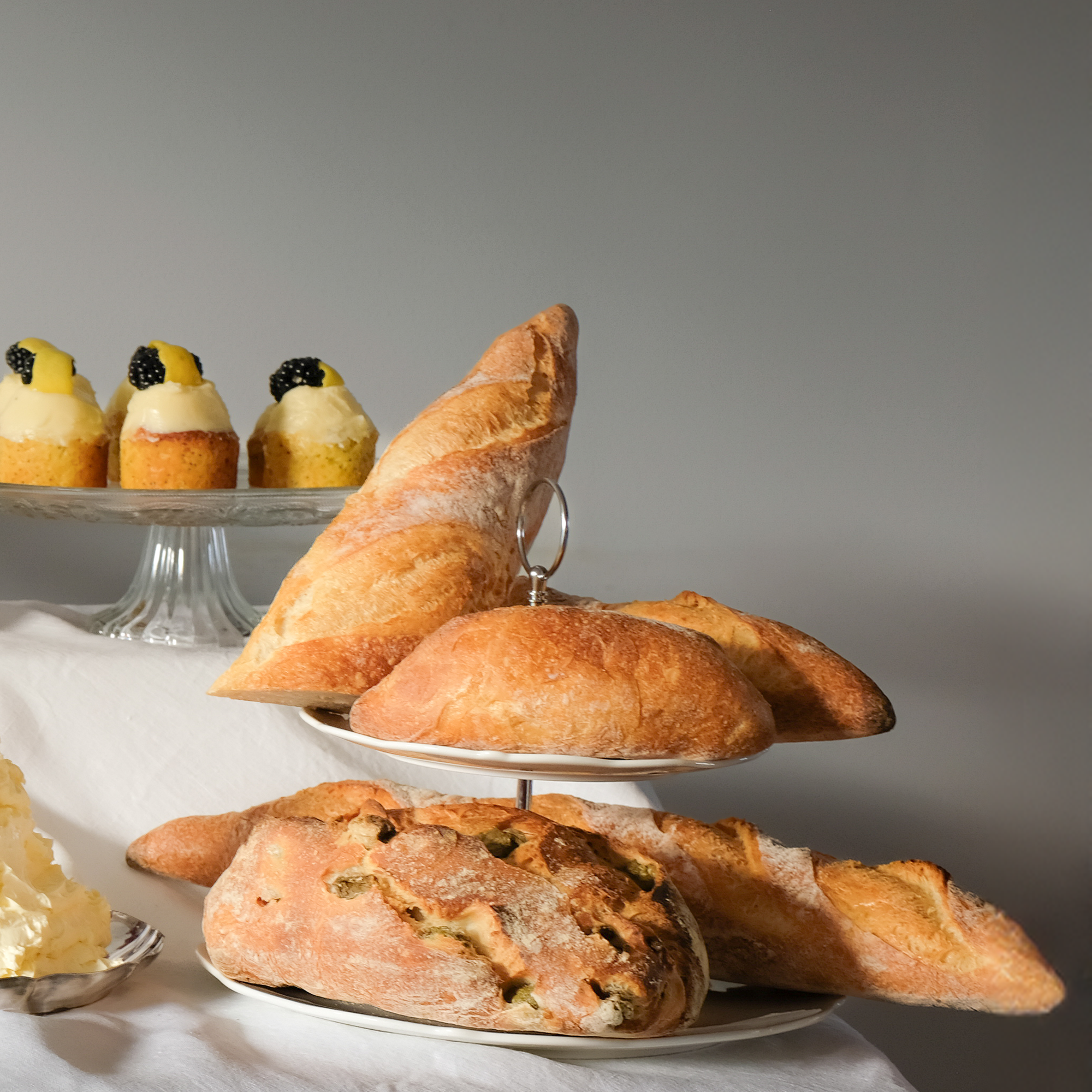 Assorted bread and pastries on white plates, with small yellow cakes topped with blackberries in the background.