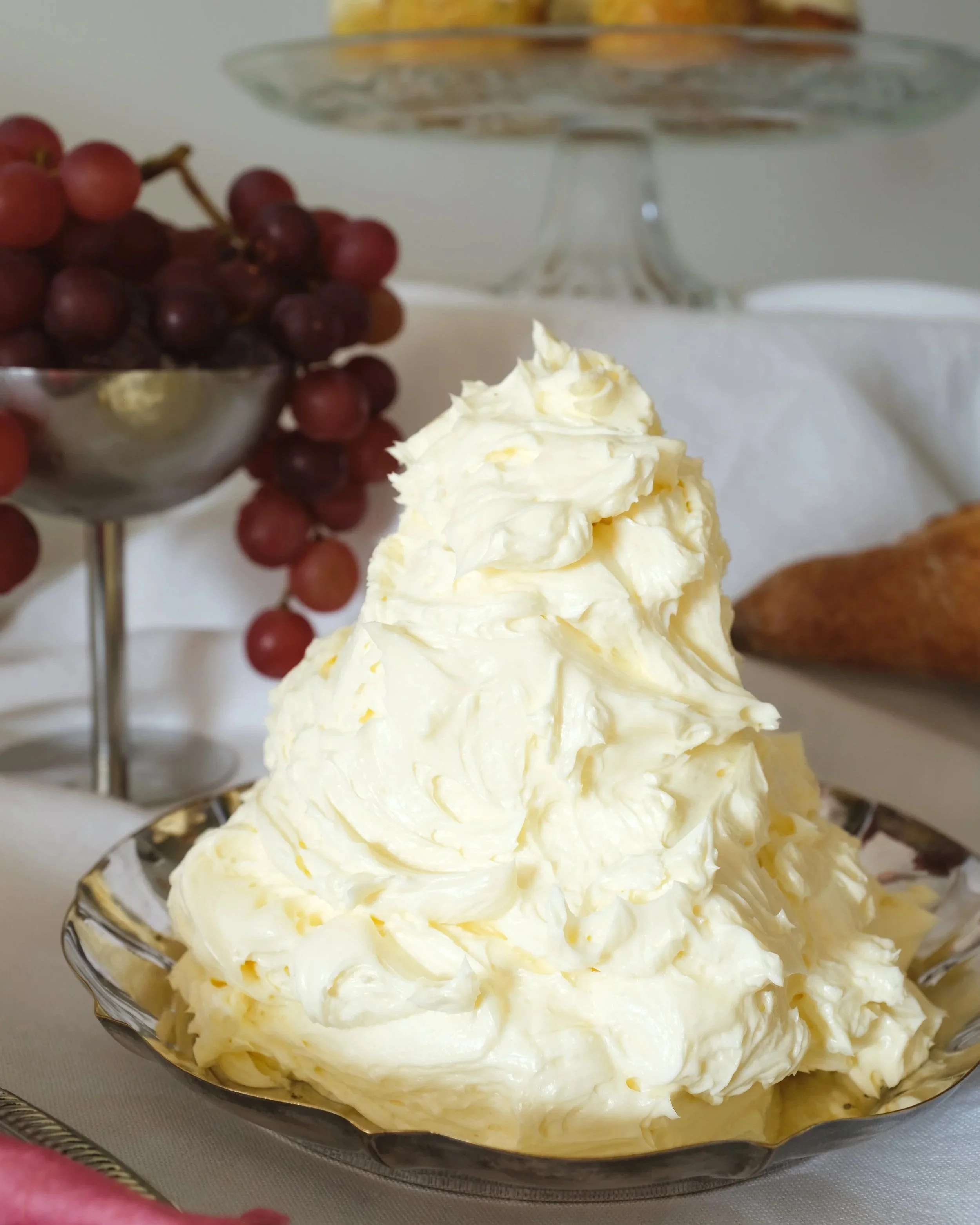 A mound of whipped butter on a glass serving dish with grapes, bread, and a cake stand in the background.