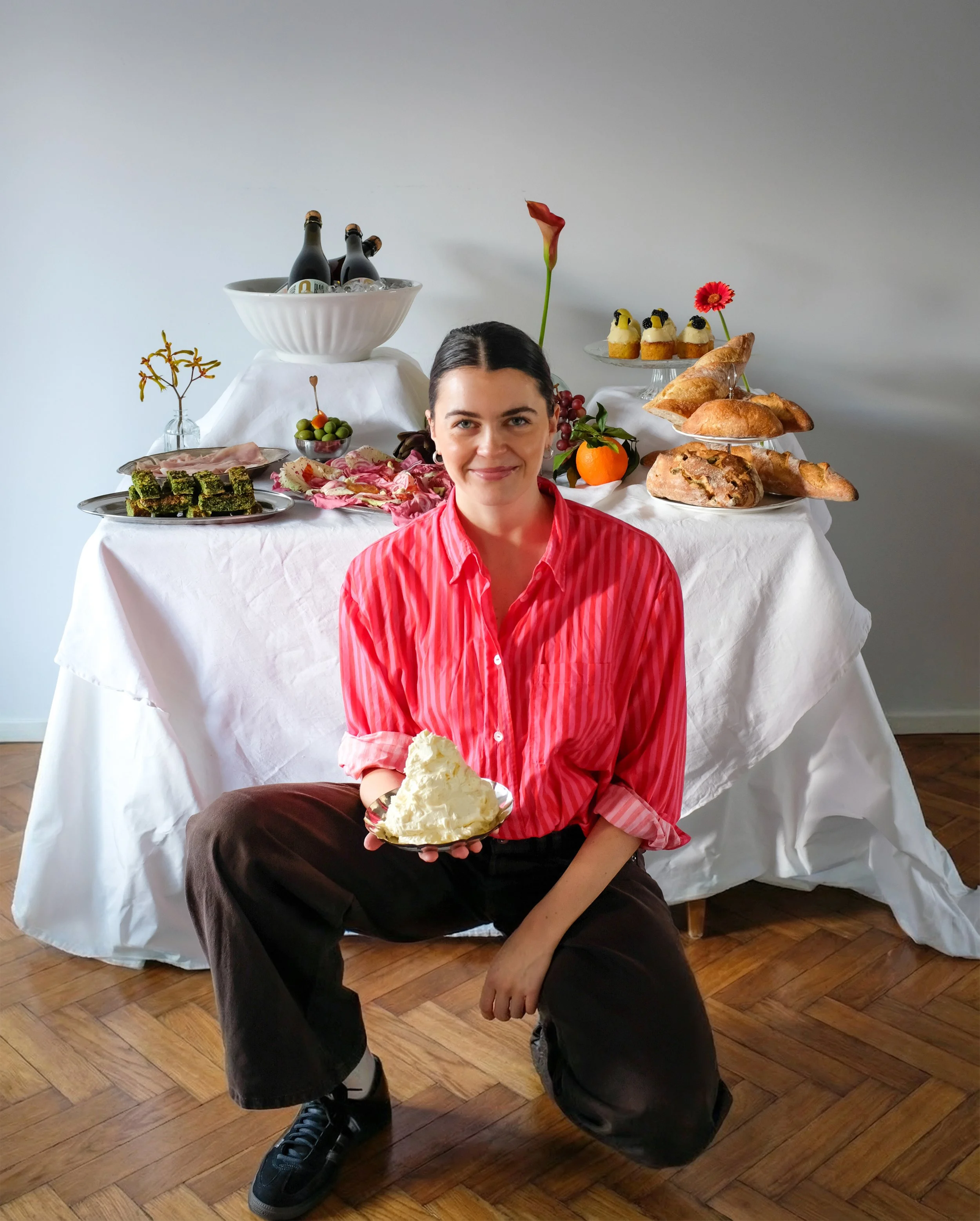 Woman sitting on the floor in front of a table with a white tablecloth, holding a slice of cheesecake. The table behind her is set with various foods including bread, cakes, and fruits, along with a bowl of champagne bottles.