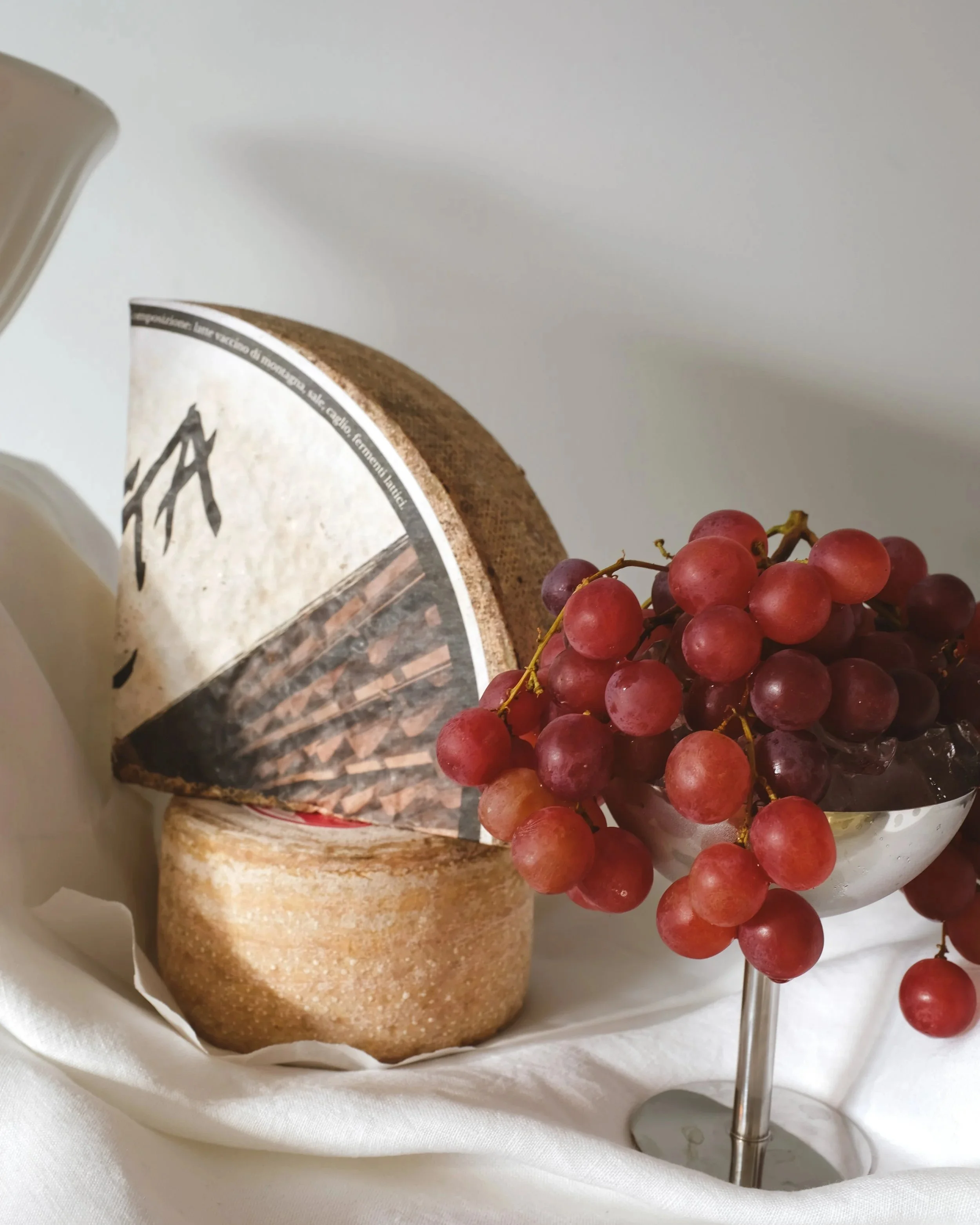 A block of cheese with a piece cut out, a bunch of red grapes in a glass bowl, set on white fabric.