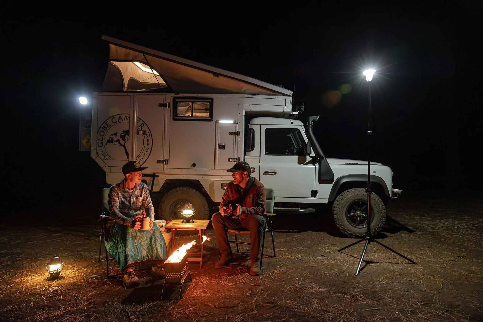 Two people sitting in camping chairs near a campfire at night, with a large camper truck behind them, illuminated by camping lights and a streetlamp.