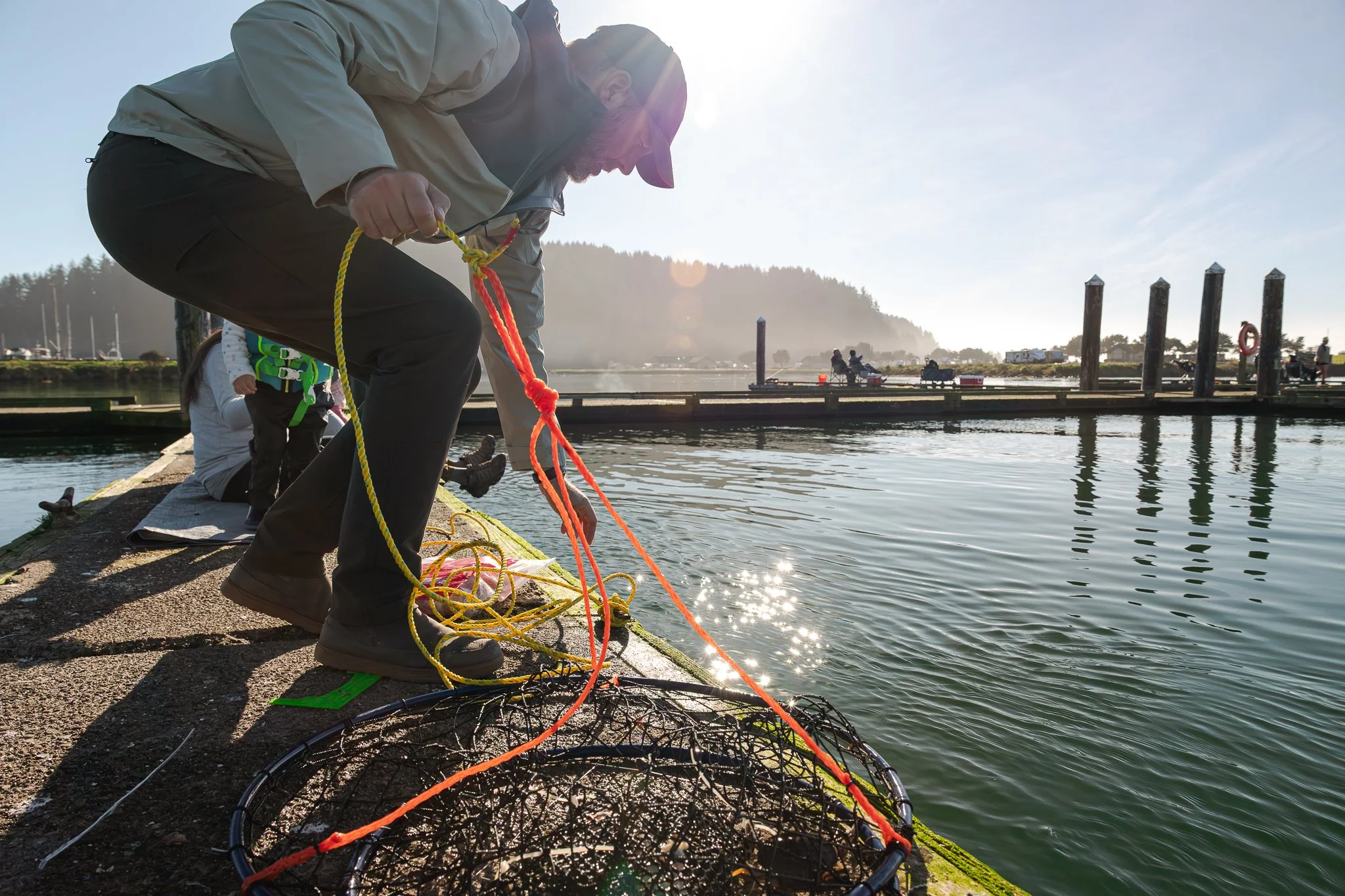 Person in Kuhl outdoor gear releasing a crab pot into the water at a pier, with a fishing net on the ground nearby, during a sunny day with a mountain in the background.