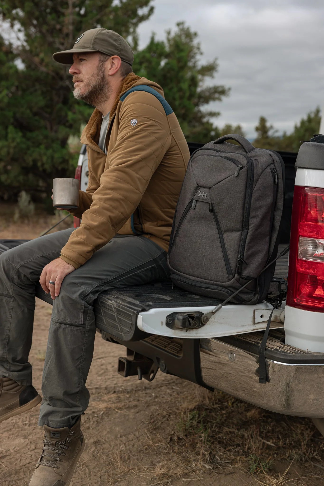 A man sitting on the tailgate of a pickup truck, holding a mug, with a gray backpack nearby, outdoors with trees and cloudy sky.