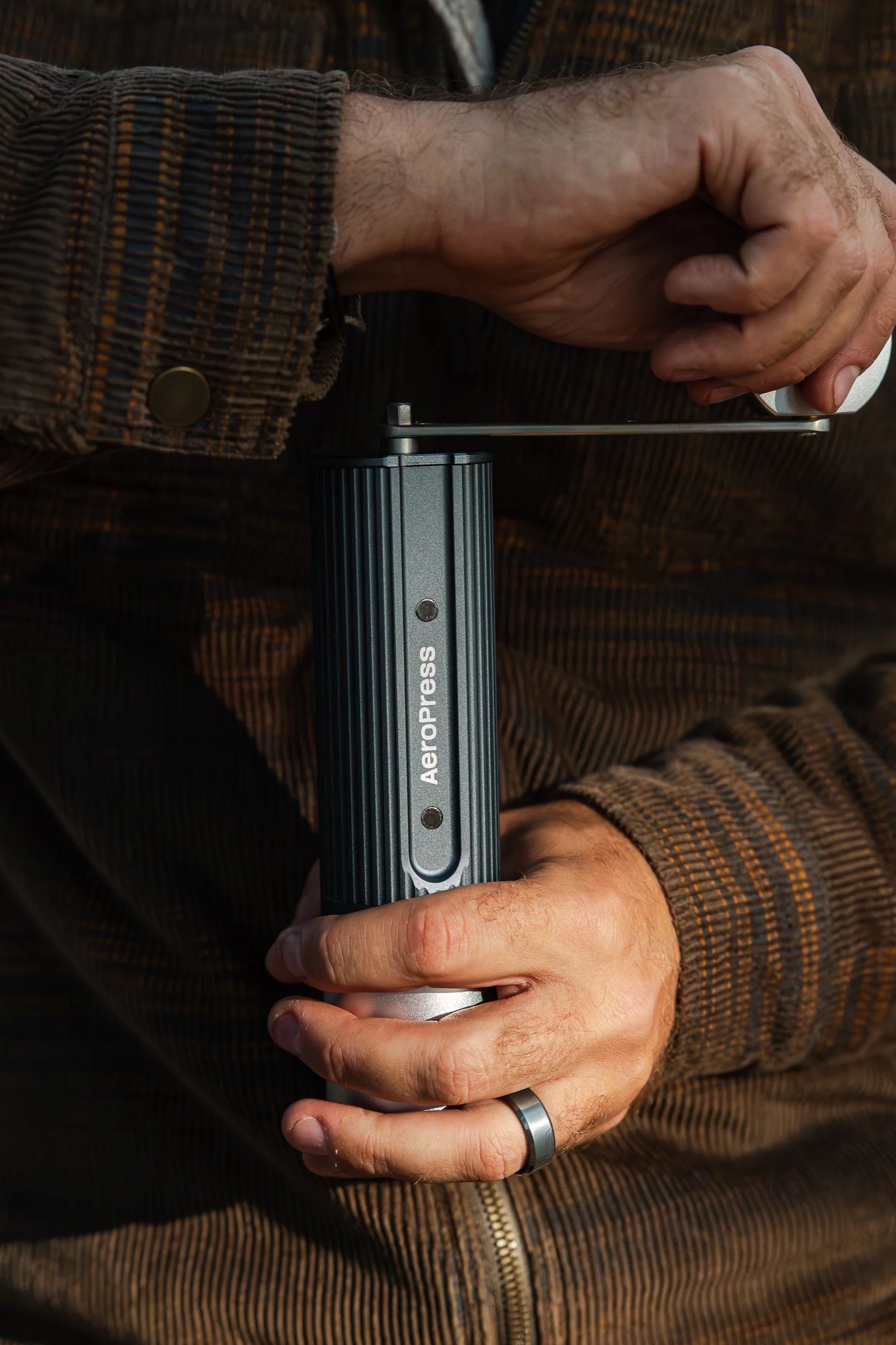 A person holding an AeroPress coffee grinder, with a focus on their hands and the device, wearing a brown striped jacket.