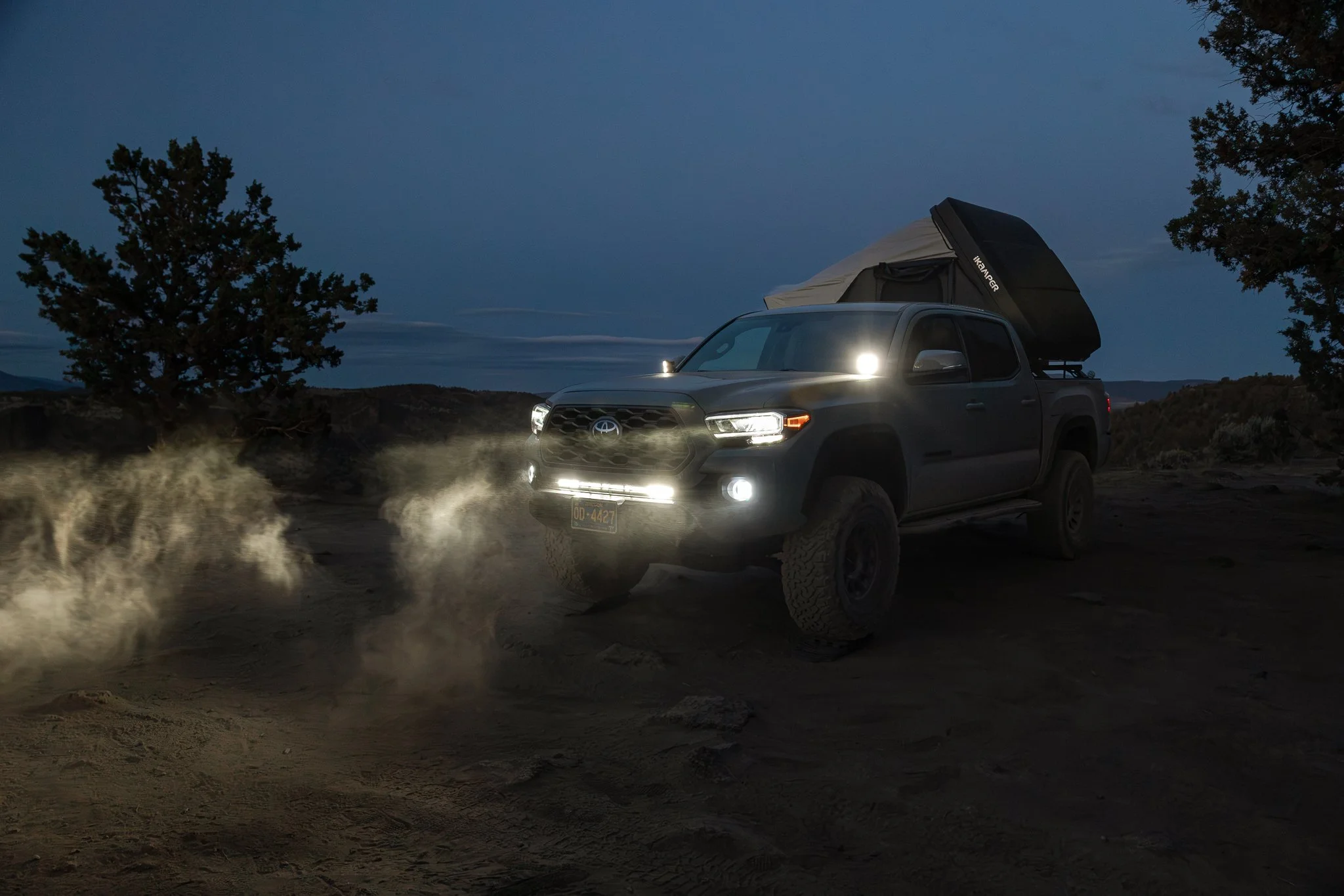 A dark pickup truck with headlights on is driving on a dirt trail at dusk or night, kicking up dust, with a tent set up in its truck bed under a dark, cloudy sky.