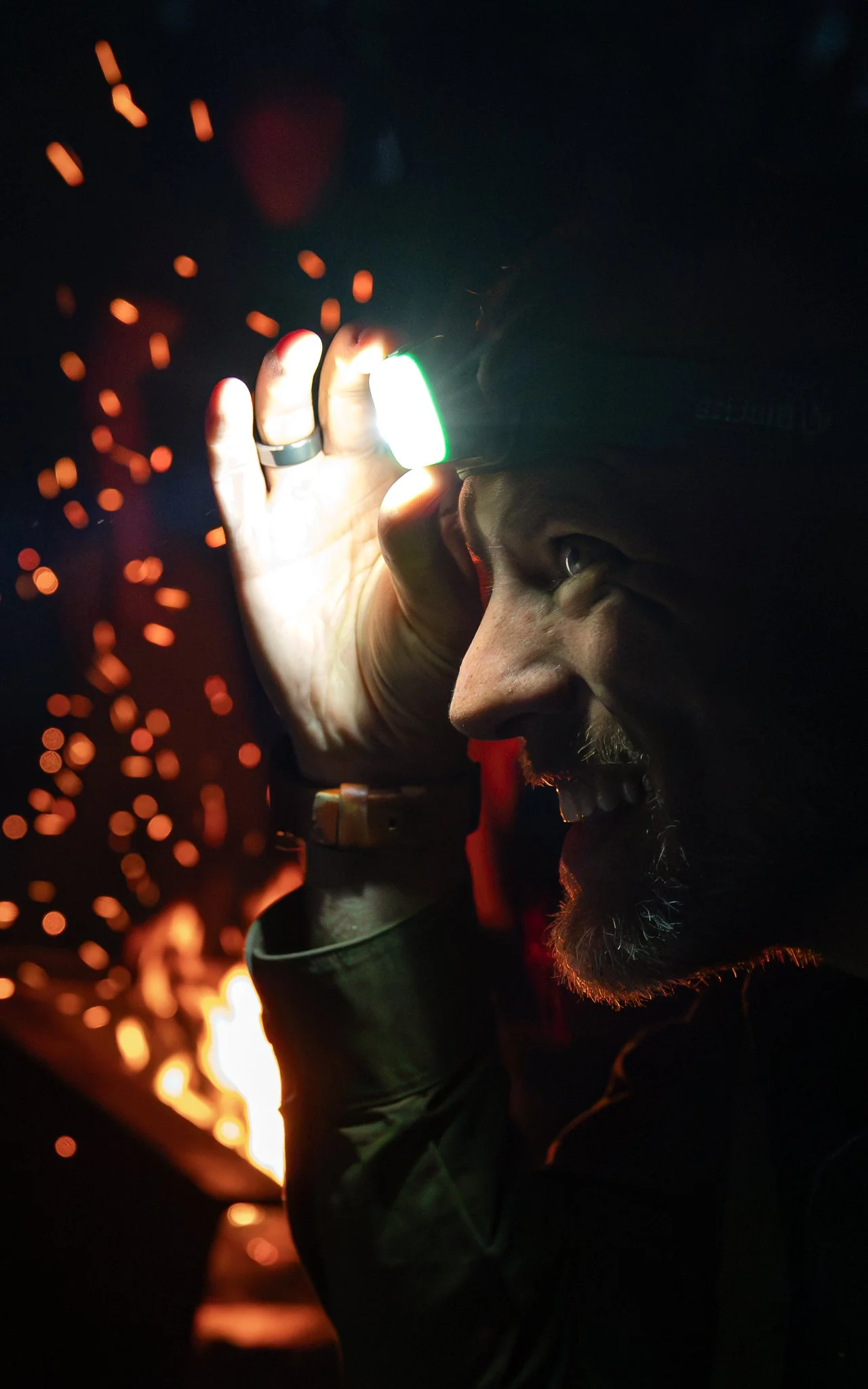 A man with a beard smiling and looking through a welding helmet, with sparks flying in the background.