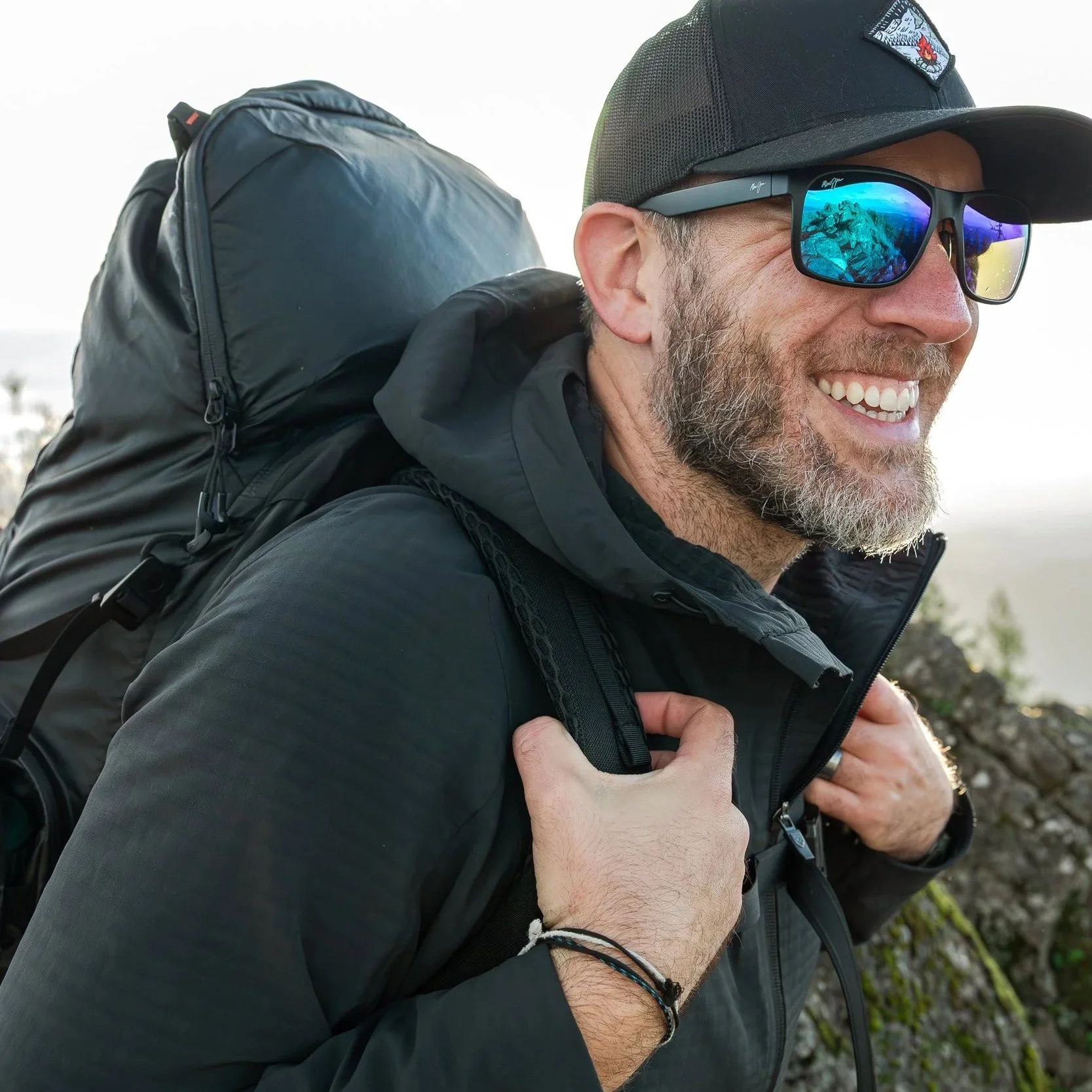 Smiling man wearing sunglasses, a black cap, and a black jacket, carrying a large backpack outdoors.