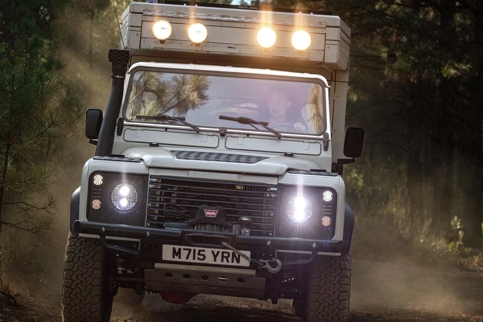 A white Land Rover Defender driving on a dirt trail in a forested area with dust in the air, headlights on, and off-road equipment and lights on the front.