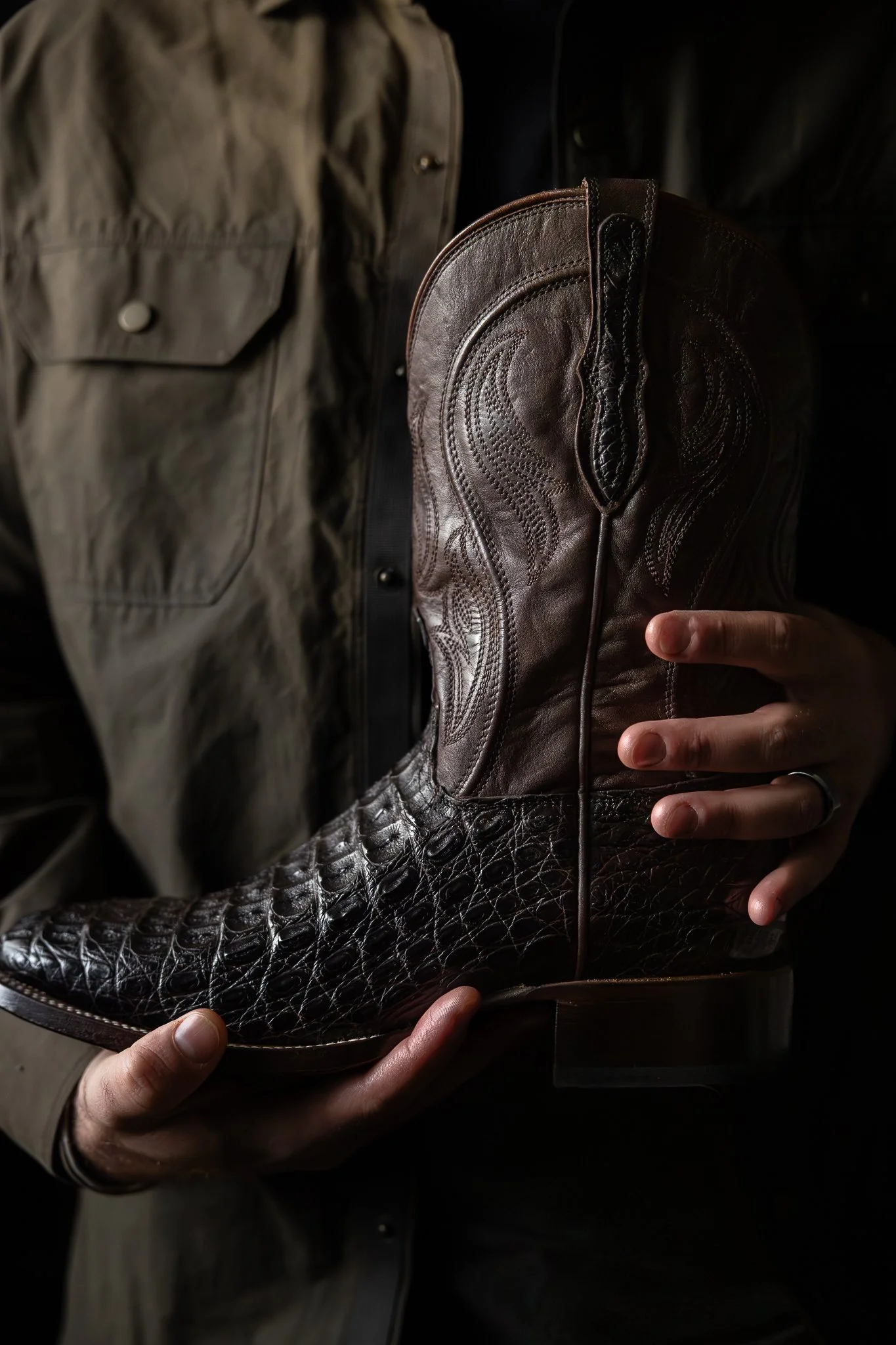 Person holding a dark leather cowboy boot with intricate stitching and a textured shaft, wearing a ring on the finger.
