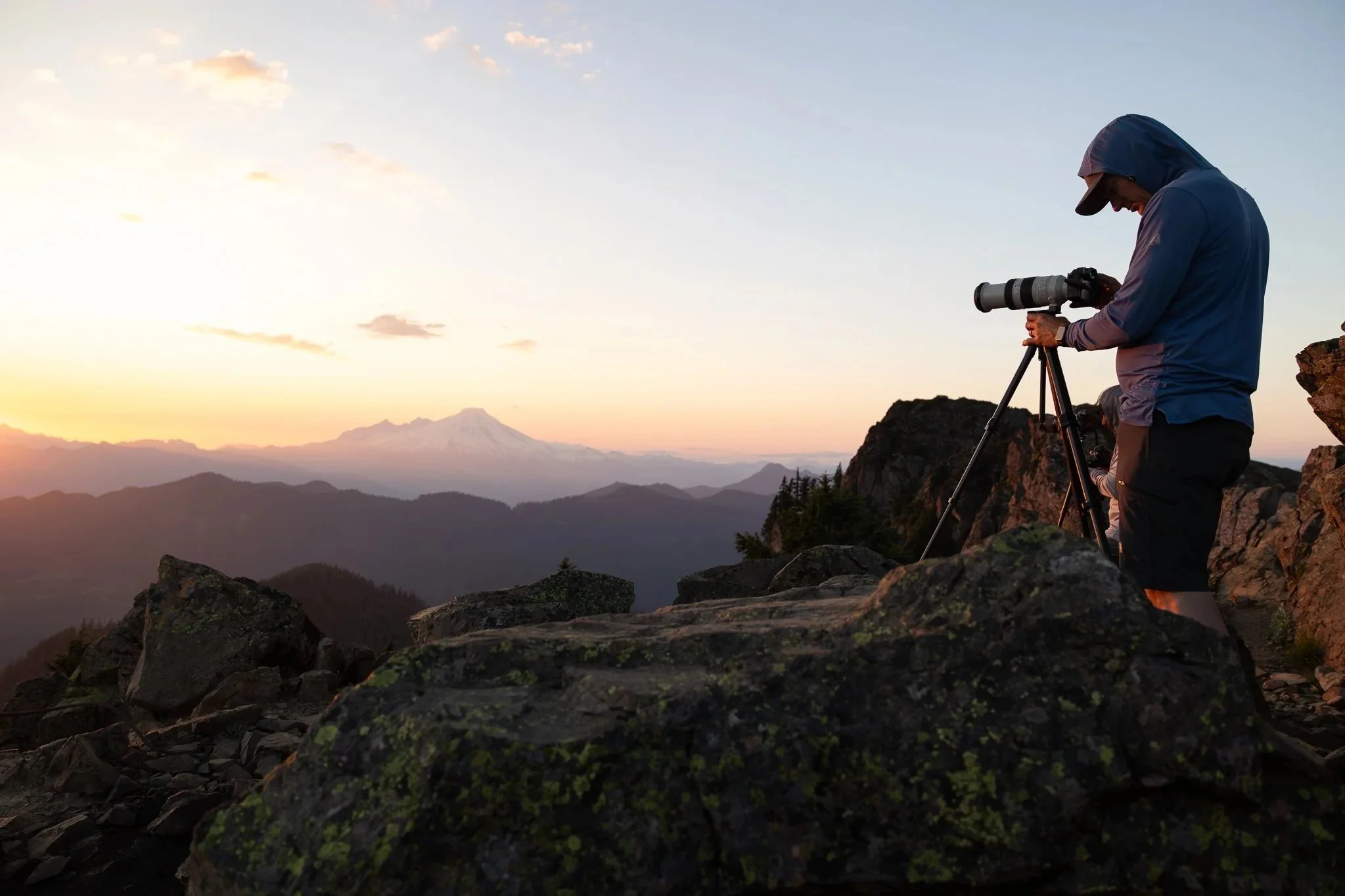 Person in a blue jacket and cap taking photographs with a camera on a tripod during sunrise or sunset in a mountainous landscape.