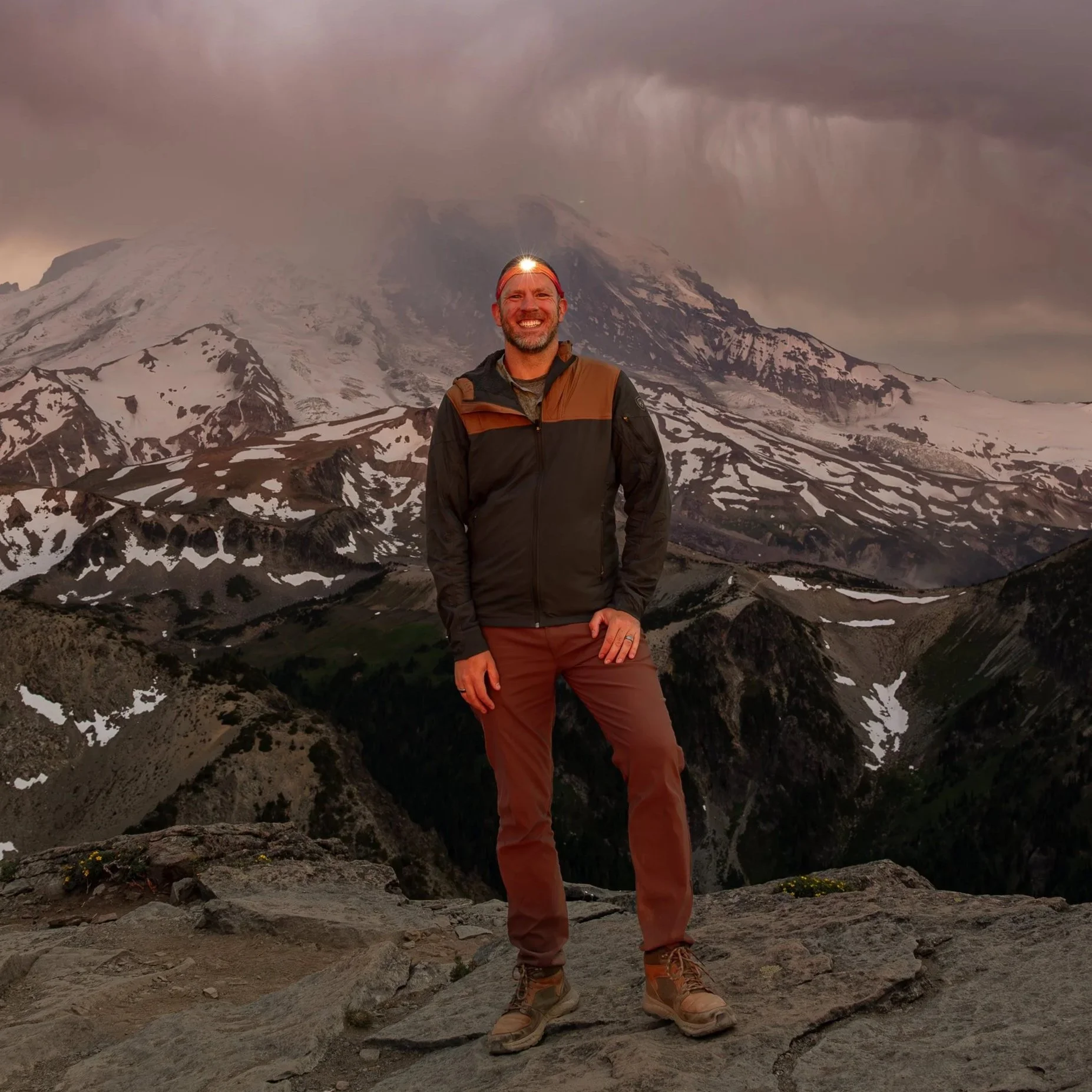A man standing on rocky terrain with snow-capped mountains and dark, cloudy sky in the background.