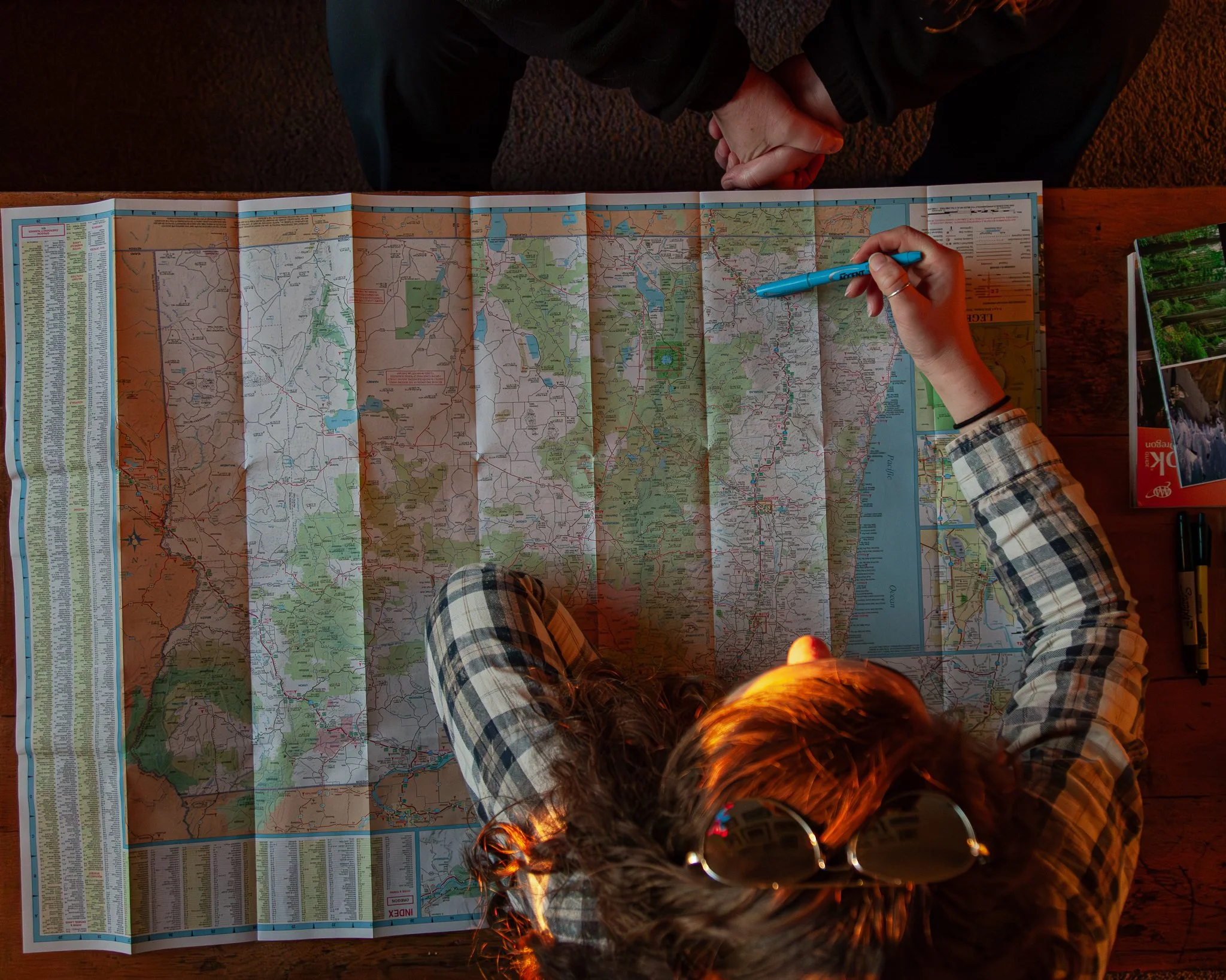 Person viewing a folded map on a wooden table, holding a blue pen, with sunglasses on their head and a magazine to the side.