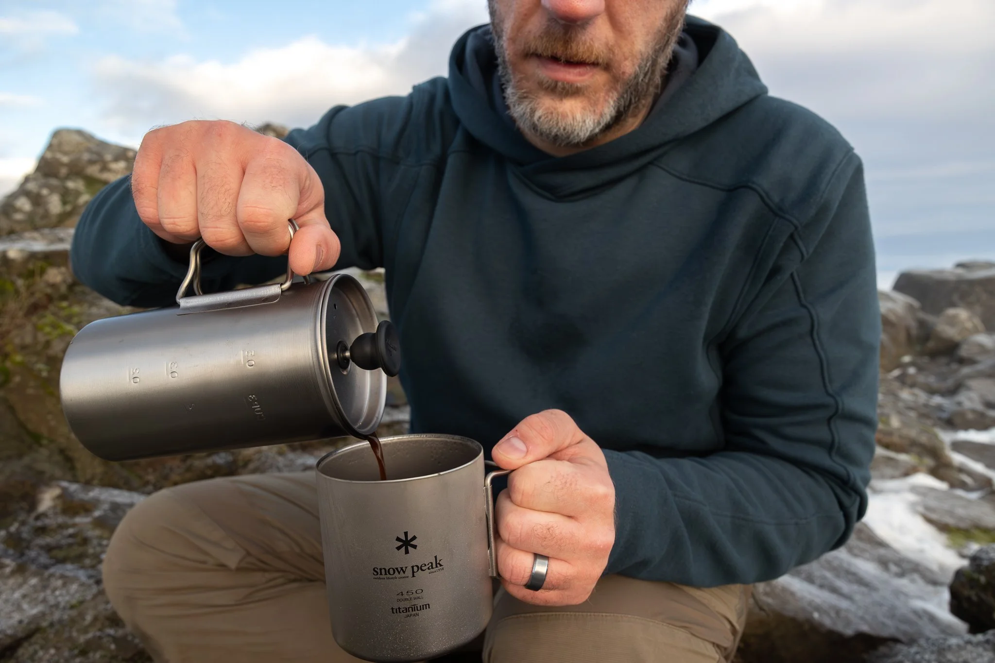 A man pouring coffee from a metal thermos into a gray mug with the Snow Peak logo, outdoors in a rocky area.