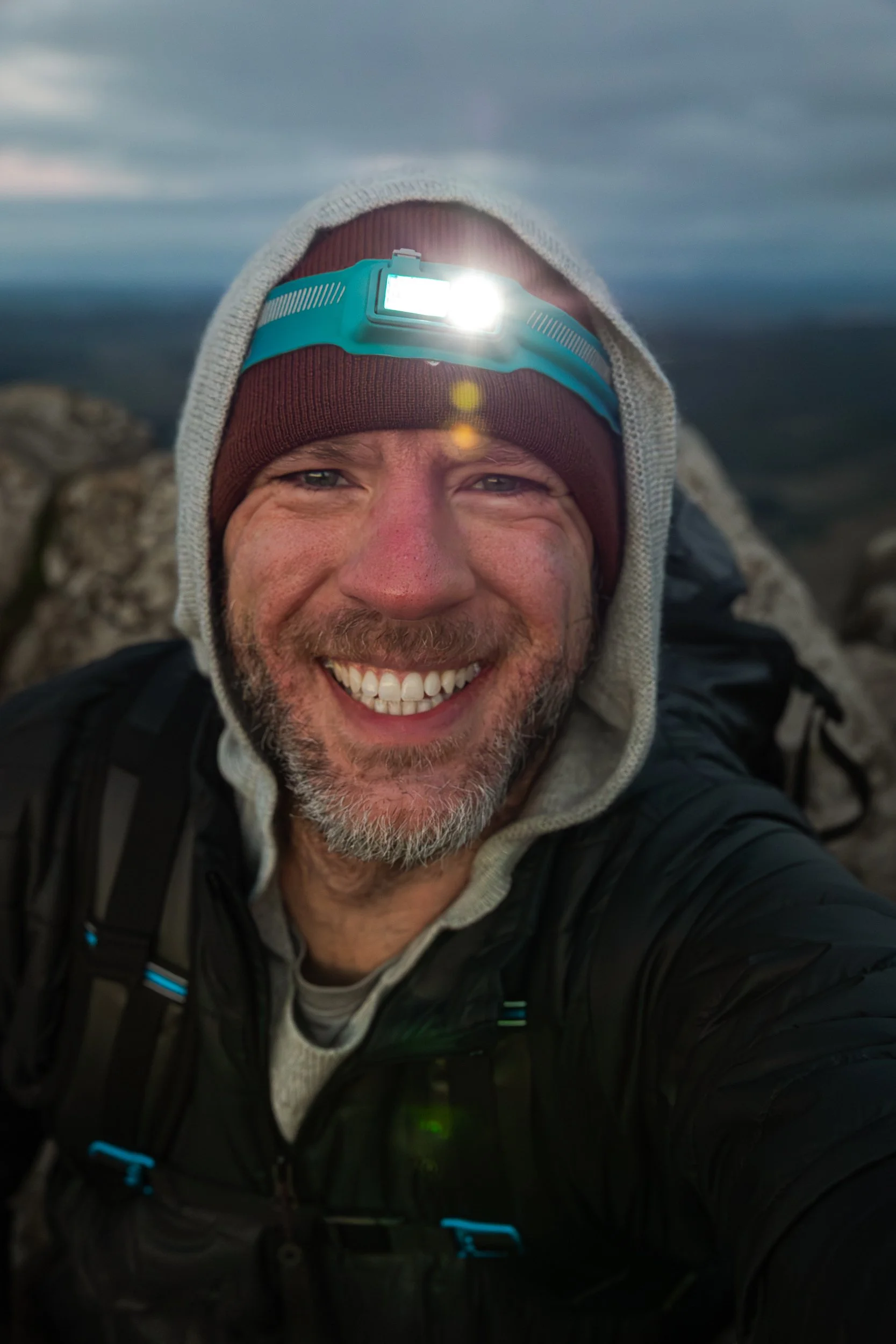 A man taking a selfie outdoors, smiling, with a mountain landscape in the background. He is wearing a headlamp, a hoodie, and a black jacket.