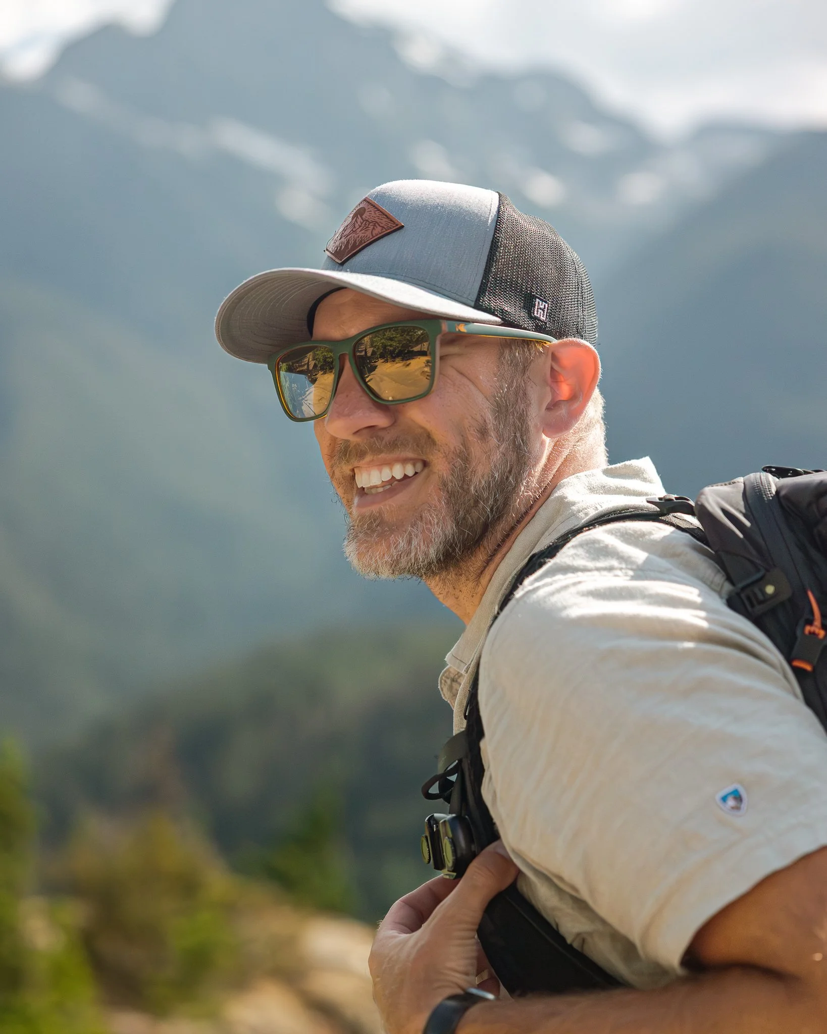 A smiling man with a beard wearing sunglasses, a baseball cap, a light-colored shirt, and a backpack, outdoors with mountains in the background.