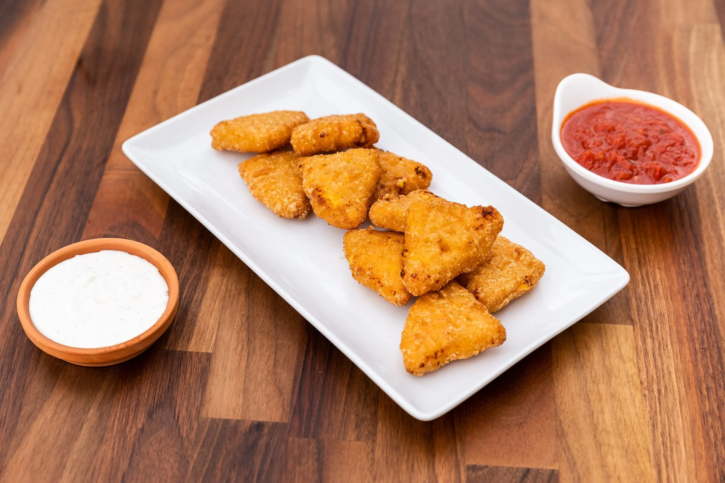 A white rectangular plate with fried fish fillets on a wooden table, accompanied by three small bowls containing tartar sauce, red salsa, and a red sauce.