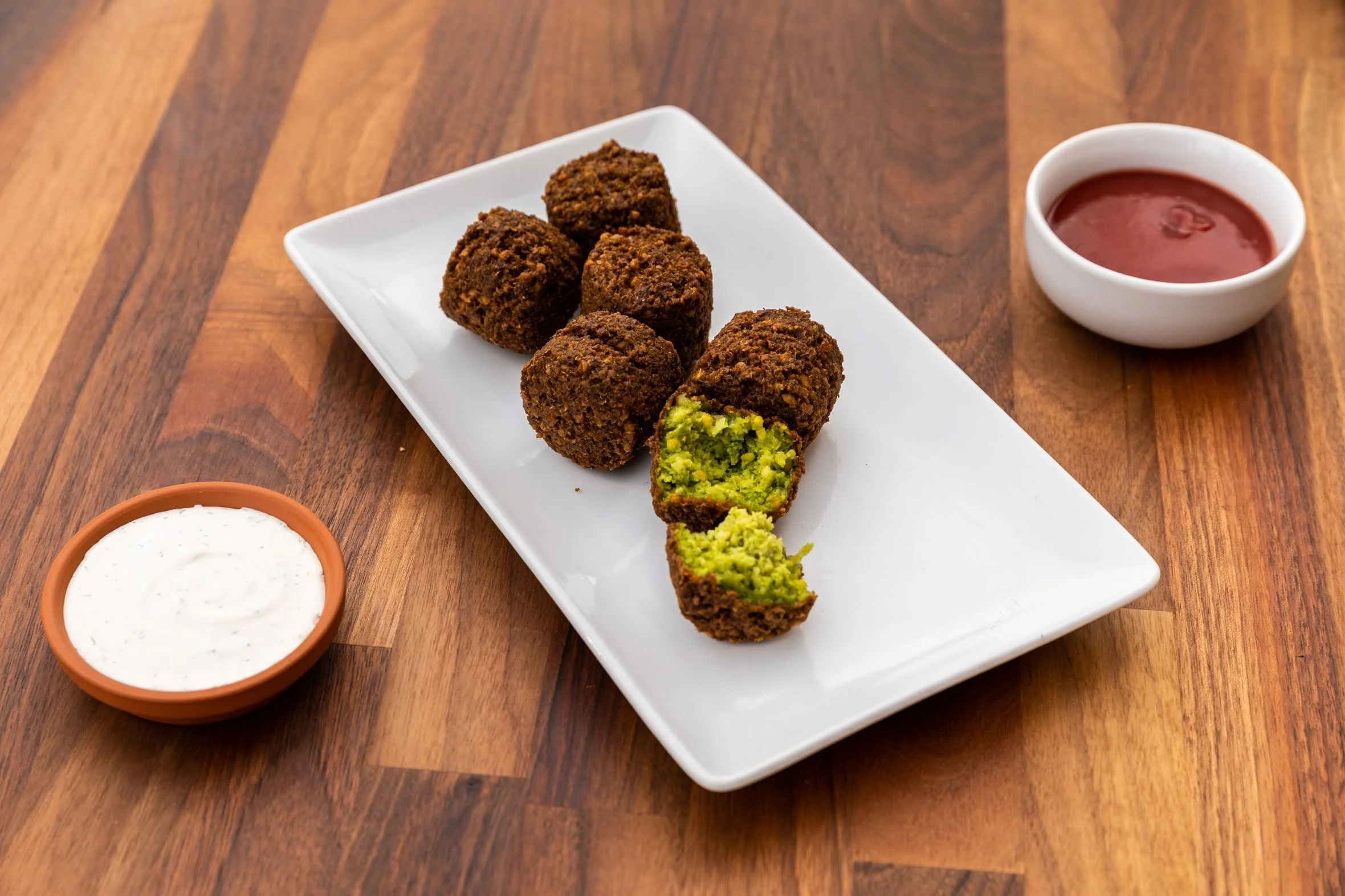 Plate of five falafel balls with one cut open showing green inside, served with sides of white yogurt sauce, red tomato sauce, and a small bowl of more white yogurt sauce.