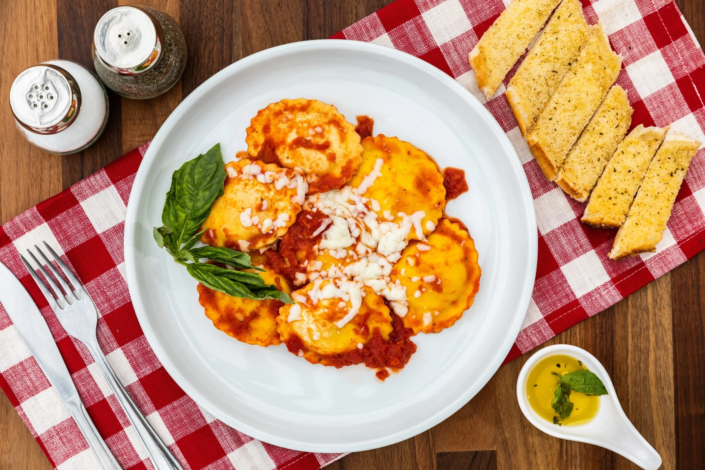 Plate of five pieces of ravioli with tomato sauce and melted cheese, garnished with fresh basil, on a white plate. To the right, garlic bread slices on a red and white checkered napkin. In the top left corner, salt and pepper shakers. At the bottom r