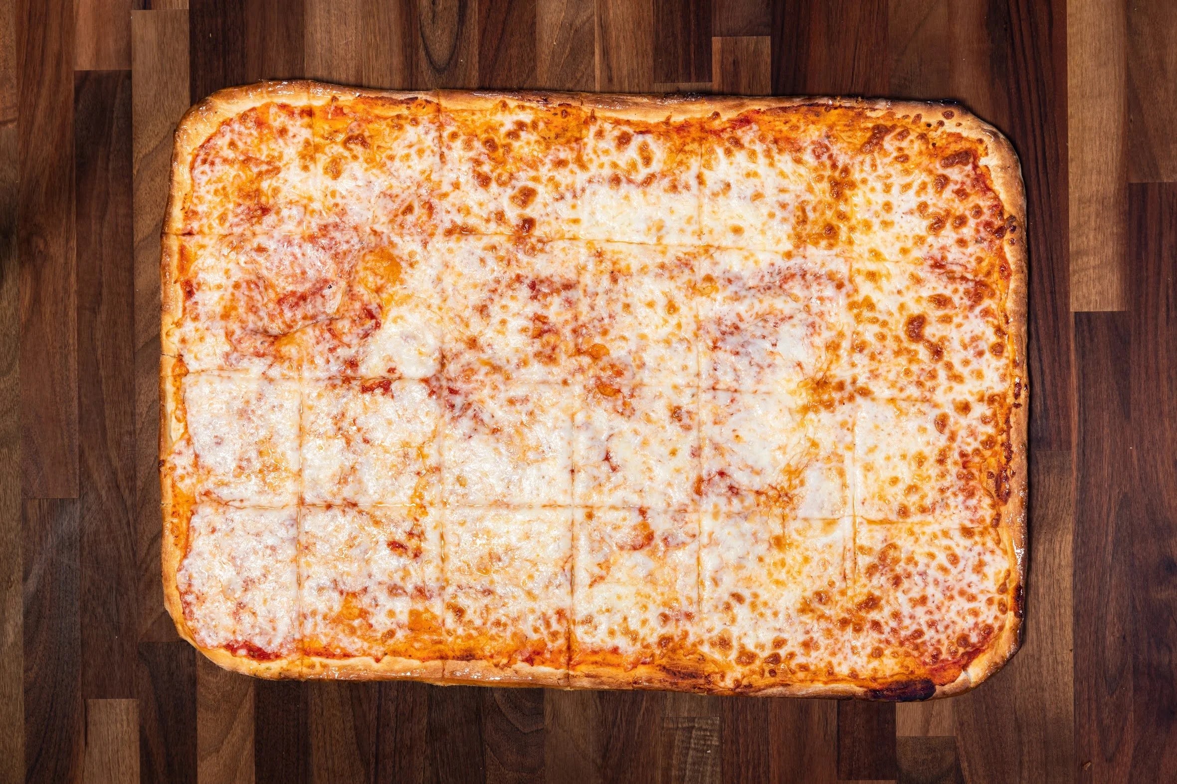 A large rectangular cheese pizza with toppings, cut into square slices, on a wooden surface.