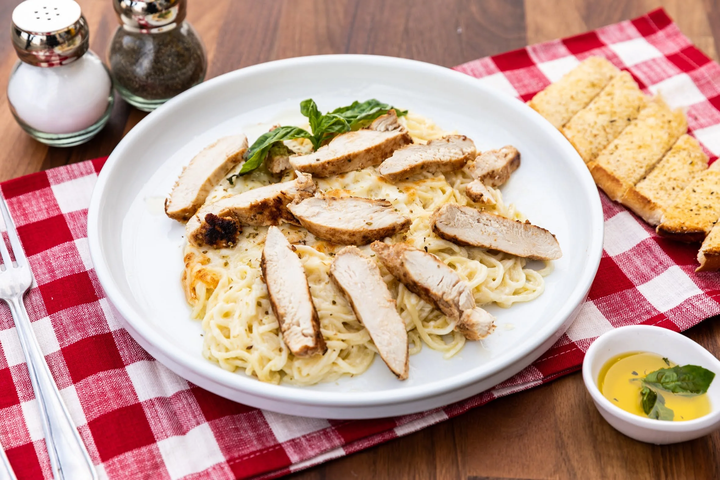 Plate of fettuccine pasta with sliced grilled chicken and basil, served on a white plate with garlic bread and condiments on a red and white checkered tablecloth.