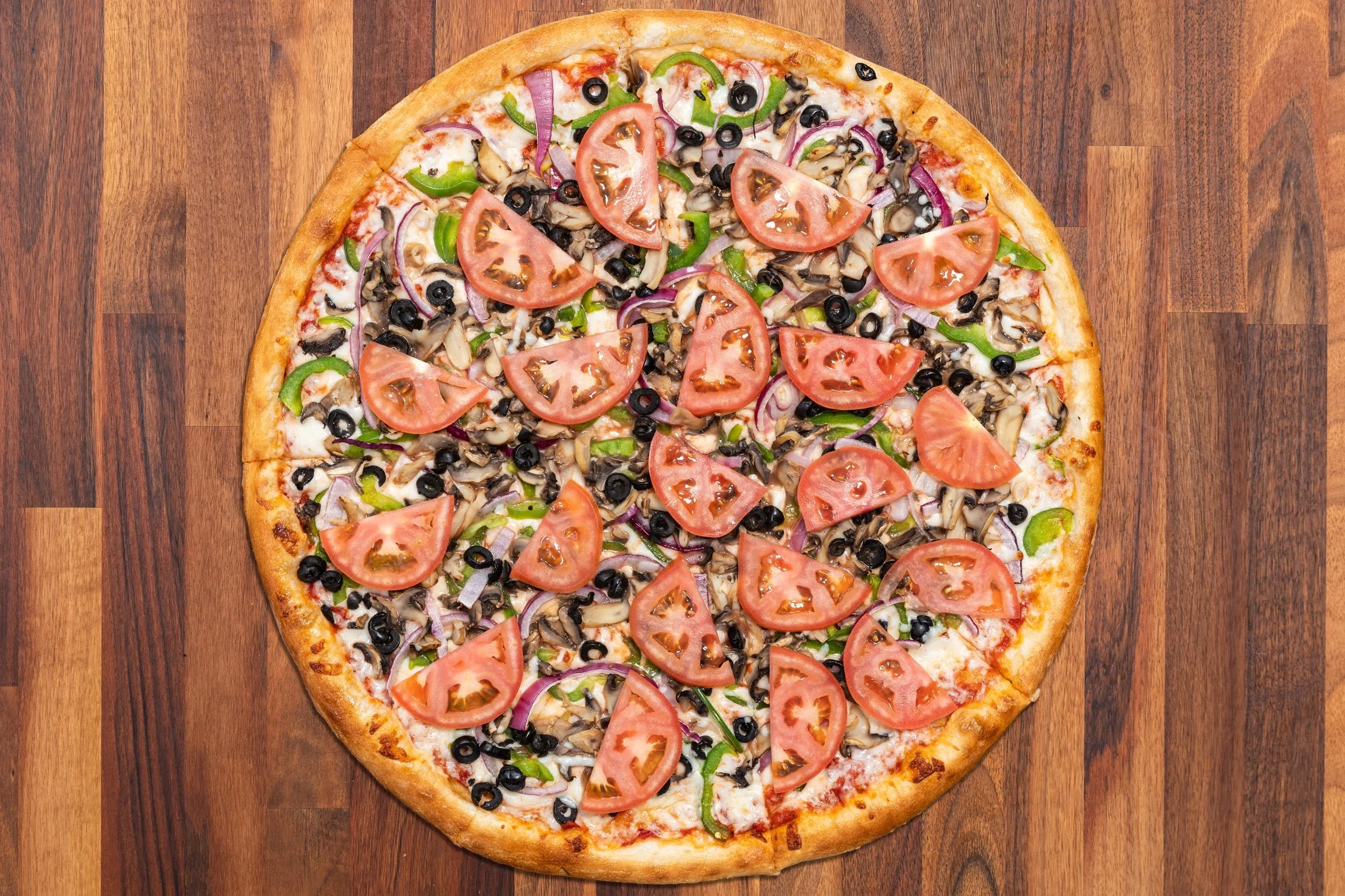 A whole pizza topped with sliced tomatoes, black olives, green bell peppers, red onions, and mushrooms on a wooden surface.