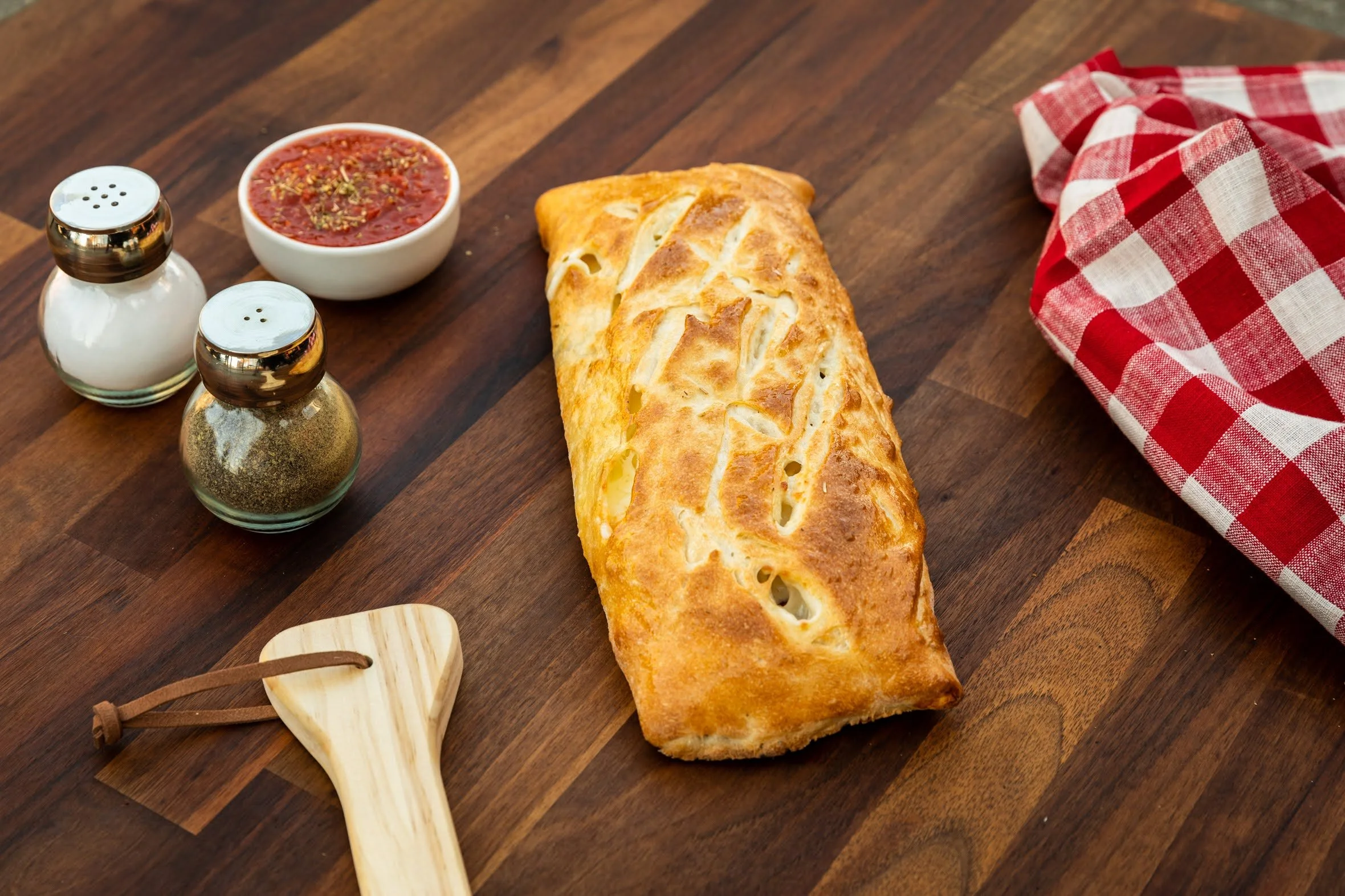 A baked calzone on a wooden table, accompanied by a small dish of marinara sauce, salt and pepper shakers, and a red and white checkered cloth.
