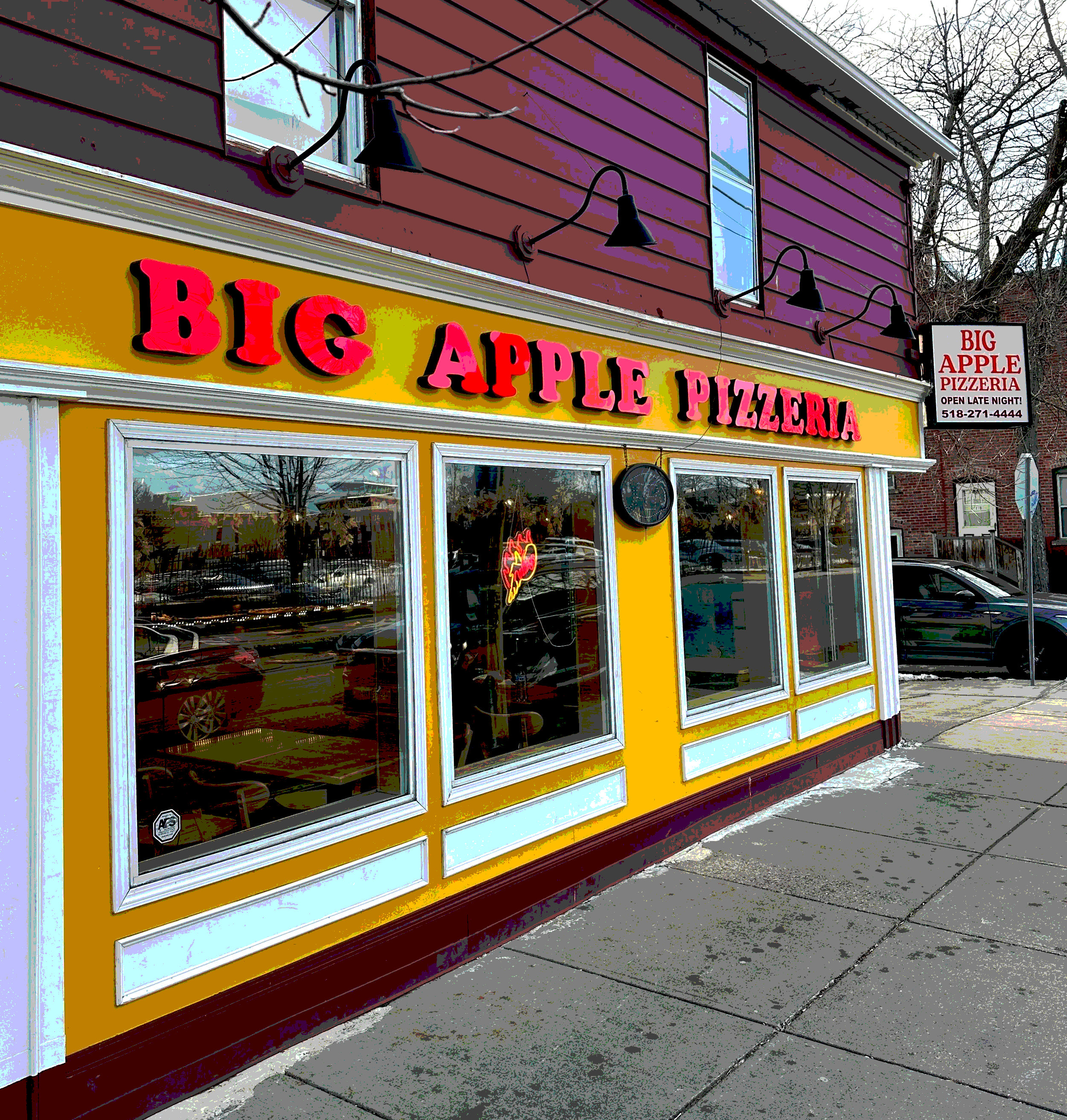Exterior of a yellow building with a sign that reads 'BIG APPLE PIZZERIA' in bright pink and red 3D letters. The windows reveal inside seating and a neon sign in the shape of a pizza slice. Above, there is a purple red-colored house with black outdoor lights and a sign indicating the pizzeria is open late with a contact number.