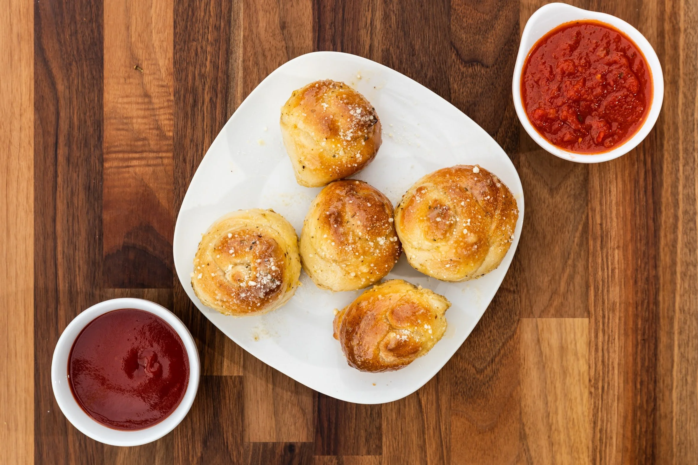 Five cheesy garlic bread rolls on a white square plate with two cups of marinara sauce on a wooden table.