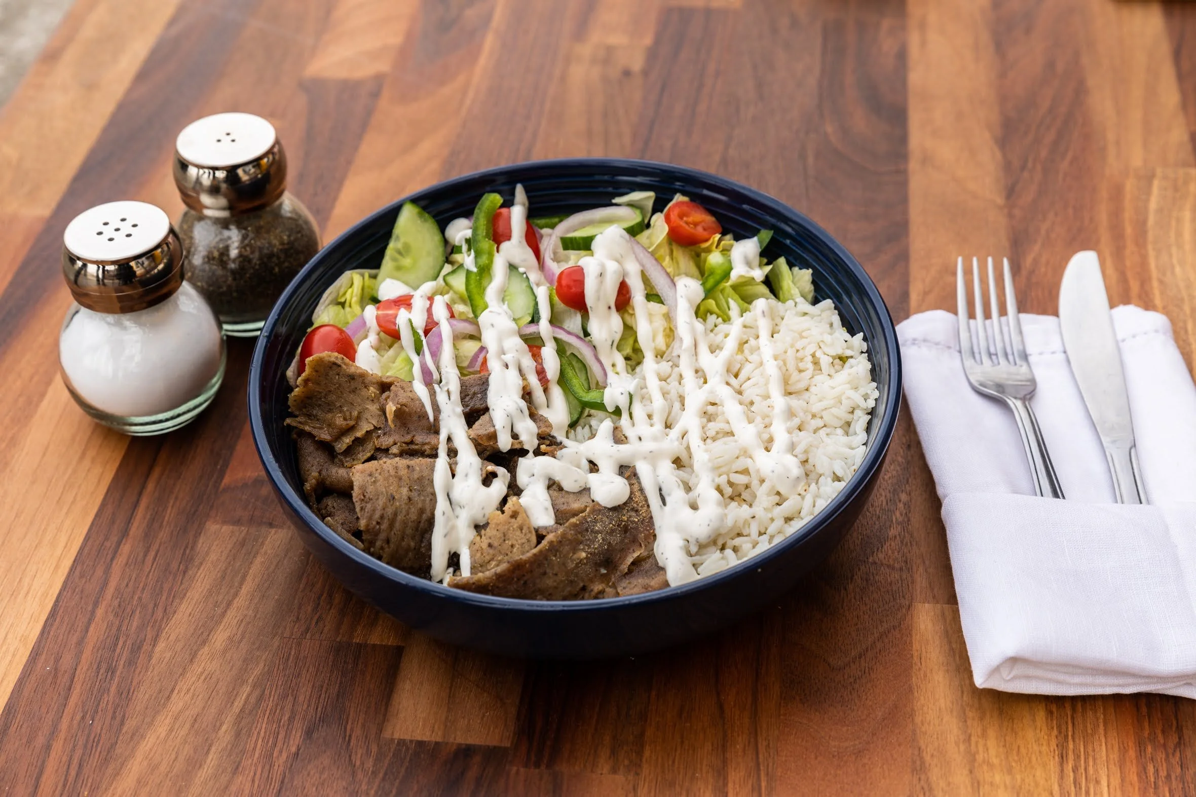 A bowl of salad with lettuce, cherry tomatoes, cucumber, and red onion, topped with shredded cheese and a drizzle of ranch dressing, served with slices of cooked beef and a side of rice, placed on a wooden table next to salt and pepper shakers and a 