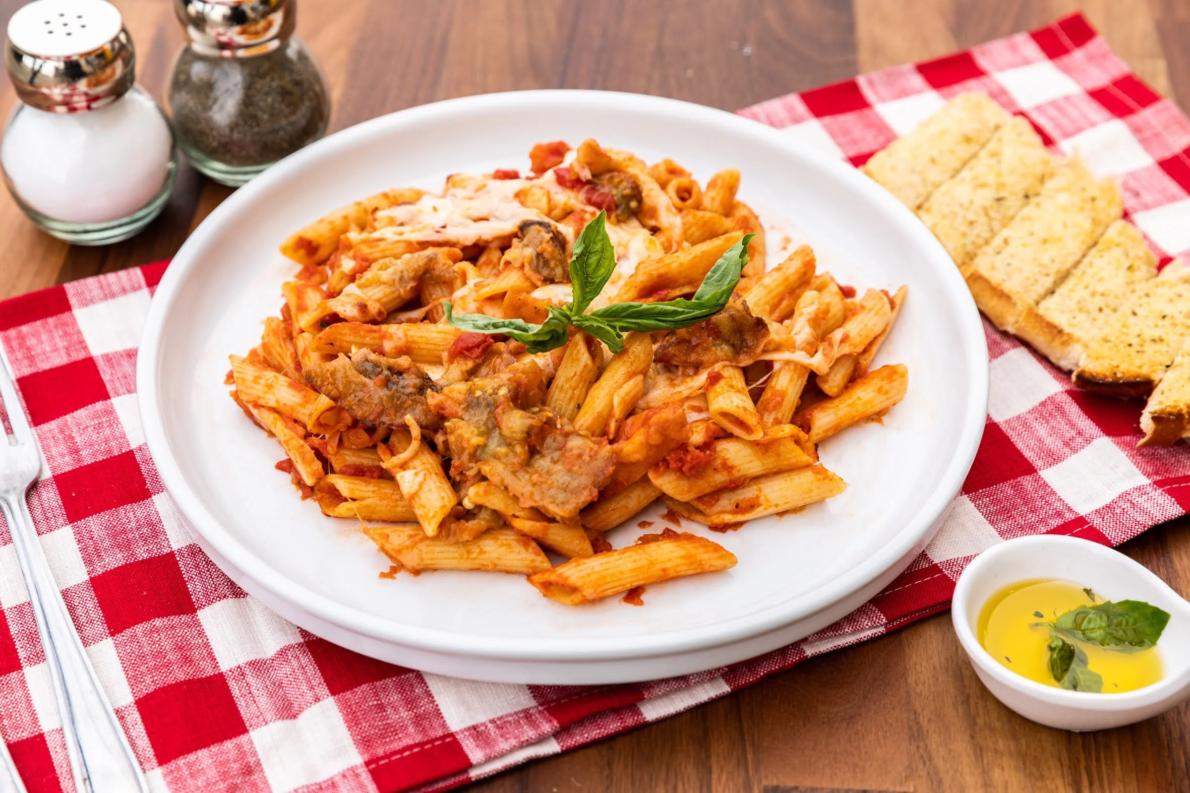 Plate of penne pasta with tomato sauce, garnished with basil, served with toasted bread, olive oil, and salt and pepper shakers.