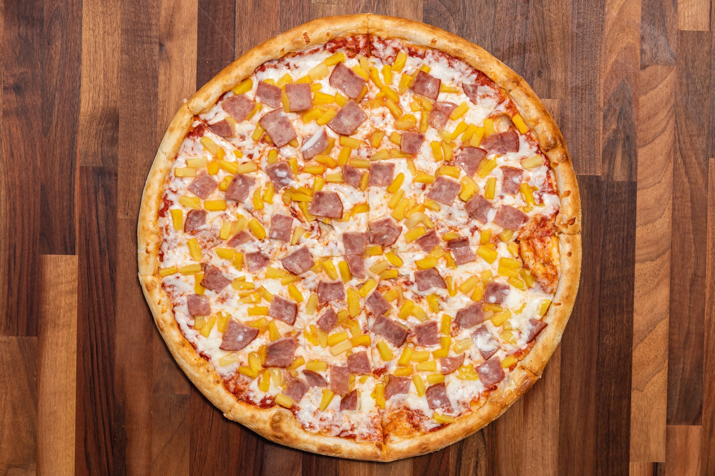 A whole pizza topped with cheese, diced ham, and pineapple slices on a wooden surface.