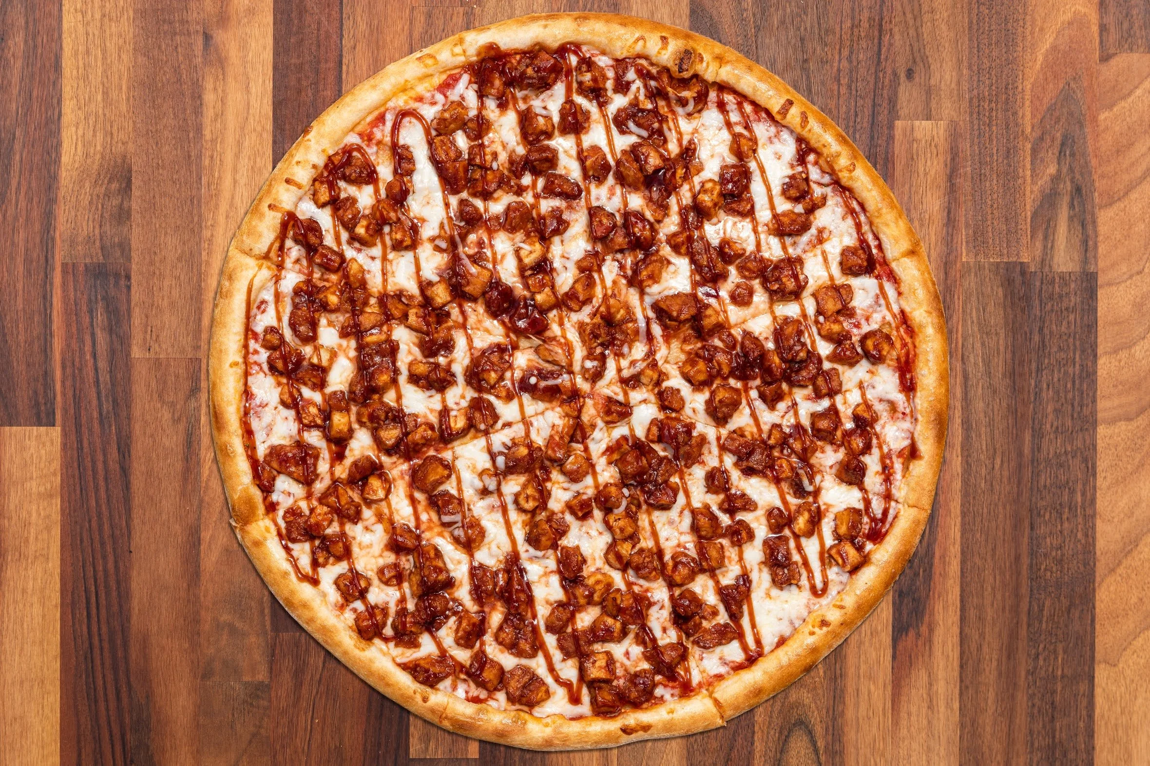 Cheeseburger pizza topped with diced bacon and drizzled with barbecue sauce, on a wooden surface.
