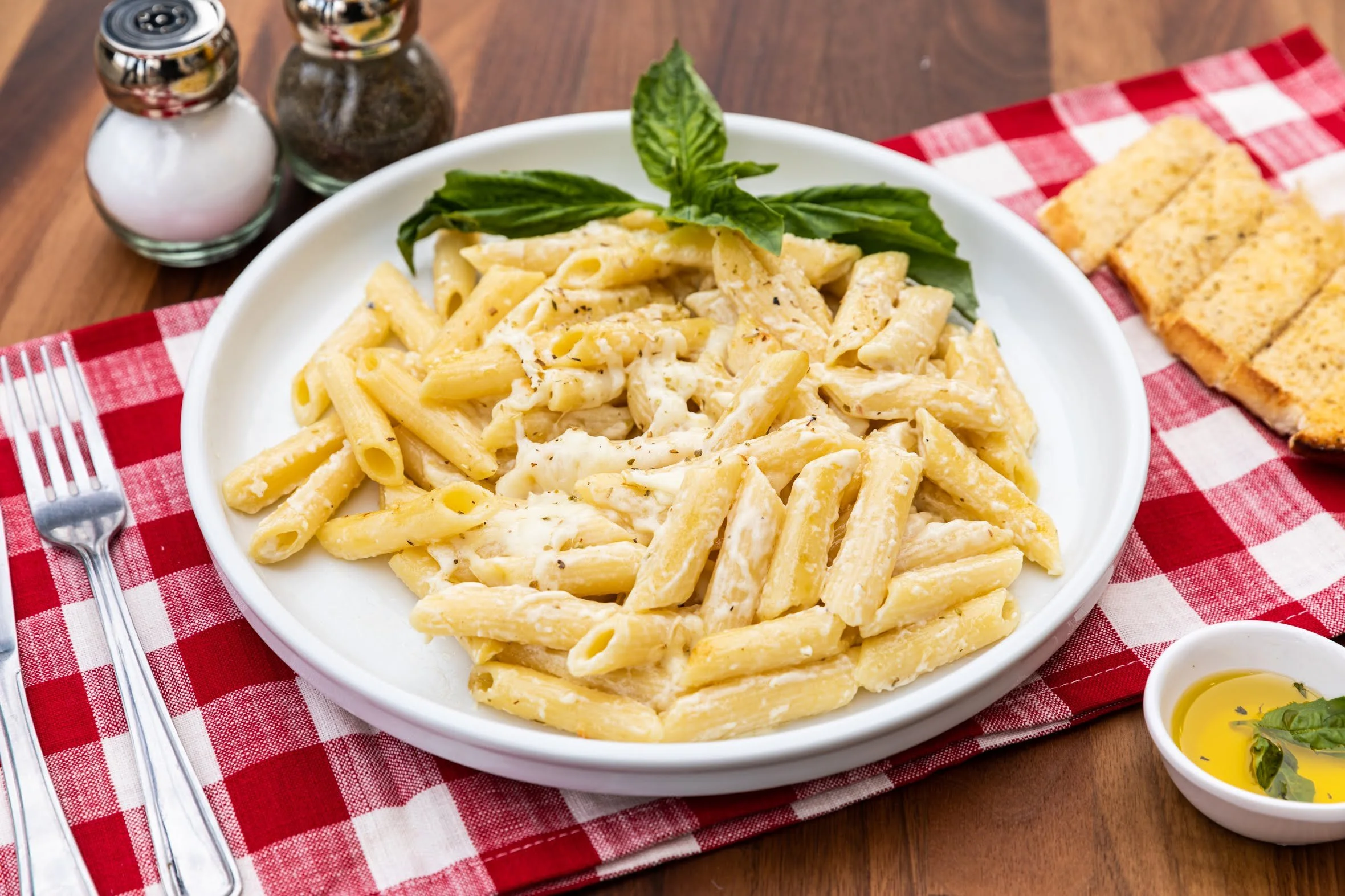 A white bowl of creamy Alfredo pasta garnished with basil leaves on a red and white checkered tablecloth with bread slices and condiments nearby.