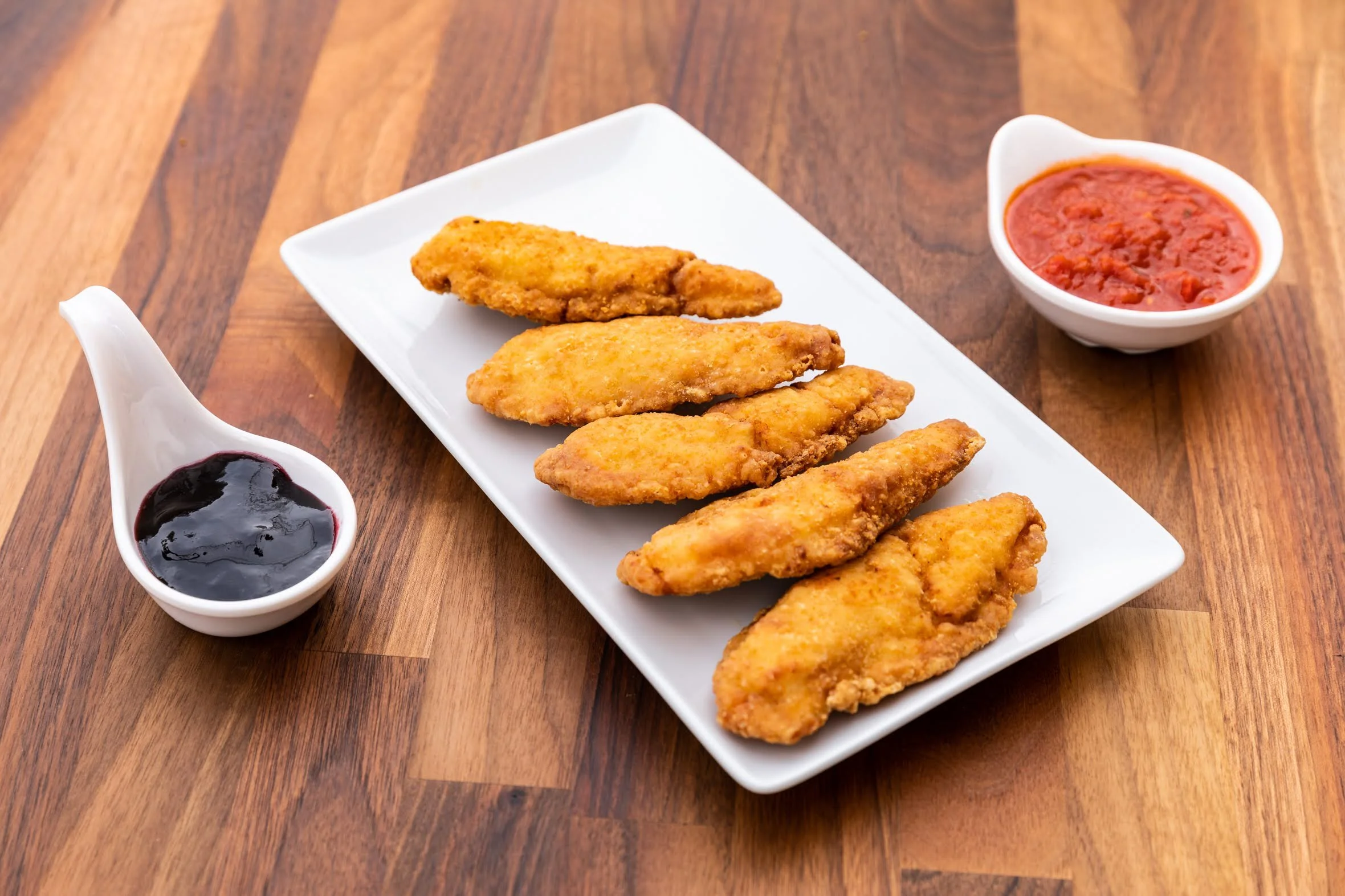 Six pieces of fried chicken tenders on a white rectangular plate, with bowls of dipping sauces on a wooden table.