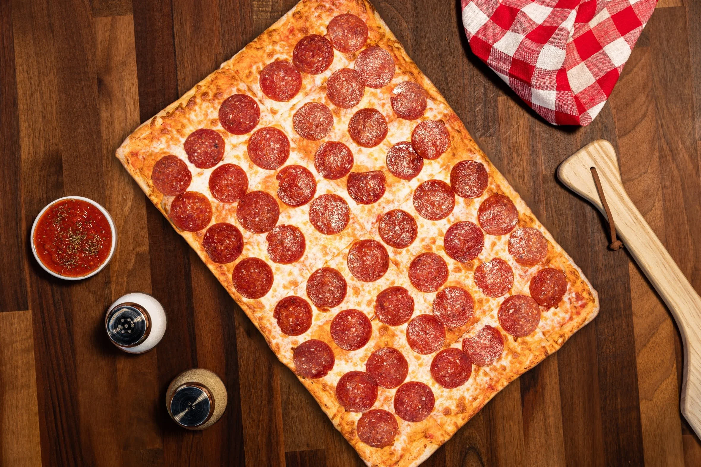 A large rectangular pepperoni pizza with cheese on a wooden table, with a bowl of marinara sauce, salt and pepper shakers, and a red and white checkered napkin.
