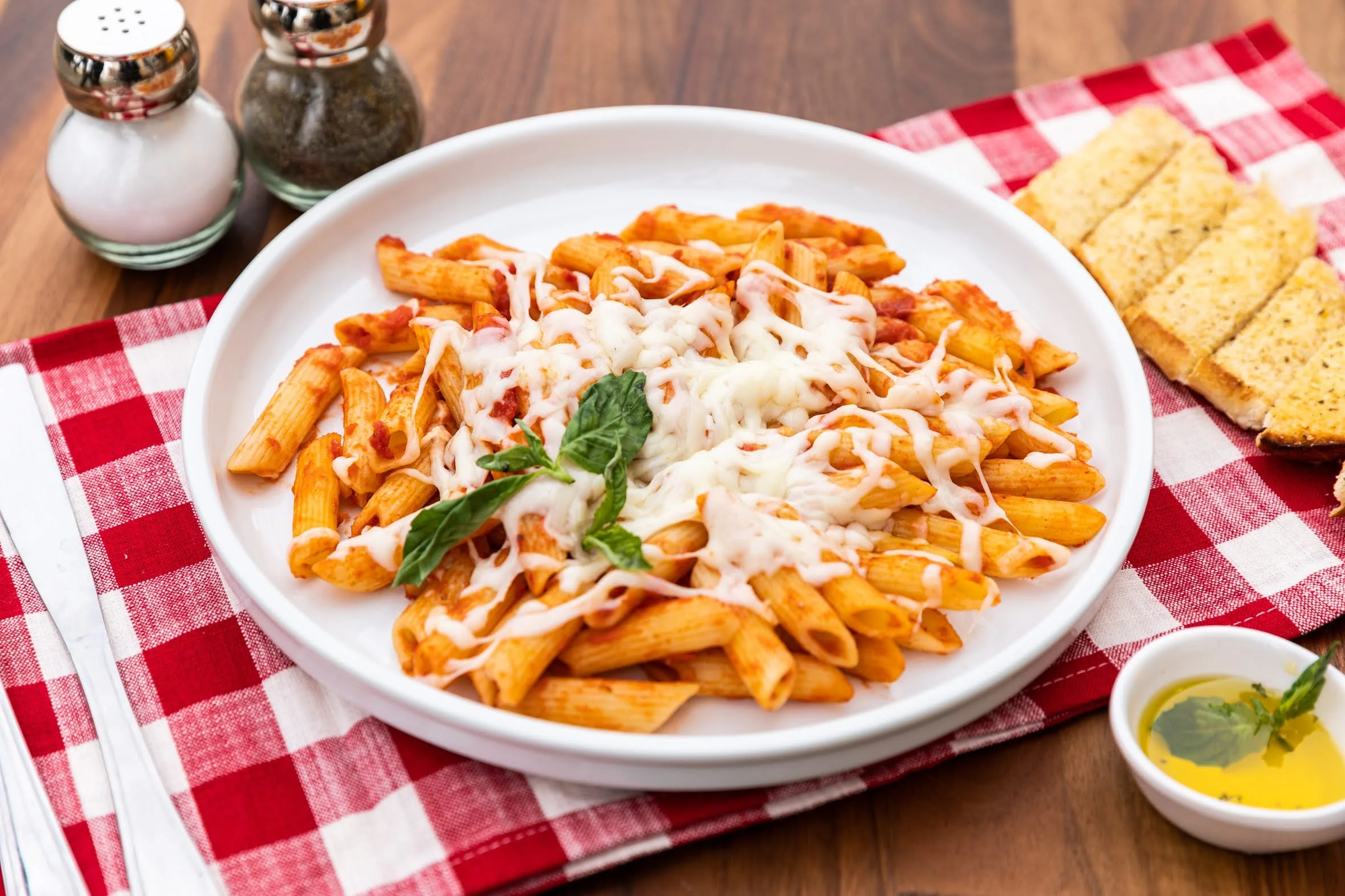 A plate of cooked penne pasta with marinara sauce, topped with melted cheese and a basil garnish, set on a red and white checkered tablecloth. Accompanying the meal are slices of garlic bread, a small bowl of olive oil with herbs, and salt and pepper