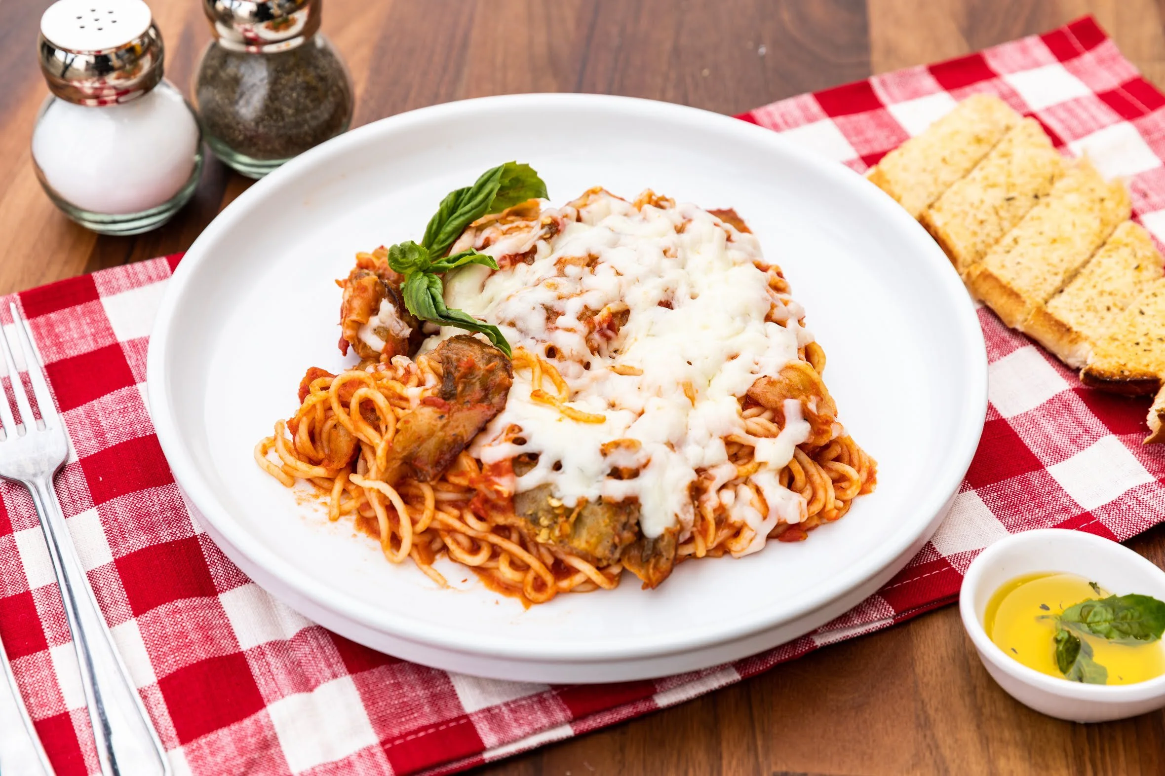 Plate of baked spaghetti with melted cheese, served with garlic bread, olive oil with herbs, and salt and pepper shakers on a wooden table with a red checkered tablecloth.