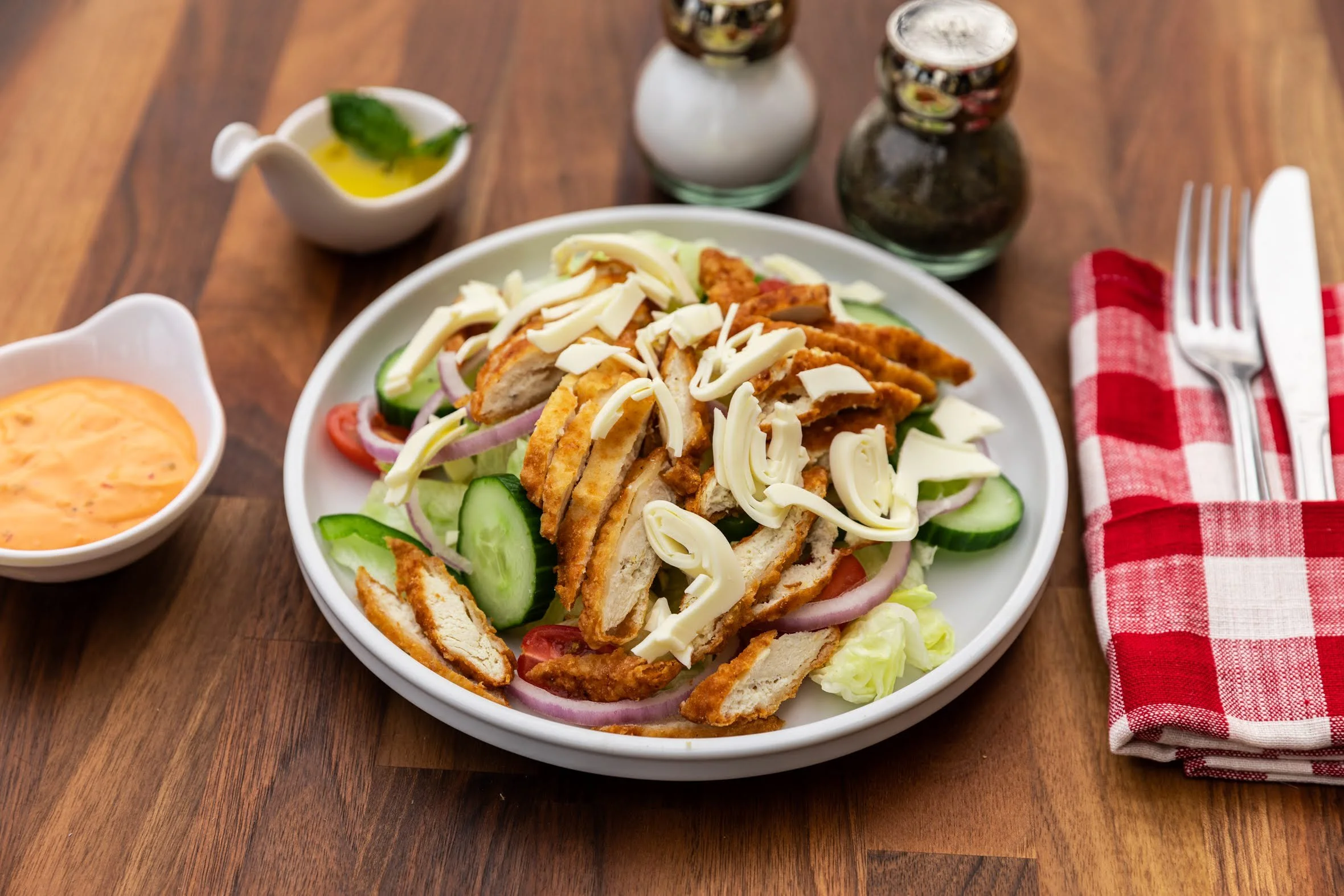 Plate of crispy chicken salad with sliced chicken, cheese, cucumber, tomato, and onion on a white dish, with salad dressing, salt and pepper shakers, and a small bowl of creamy dressing nearby on a wooden table, with utensils on a red and white check