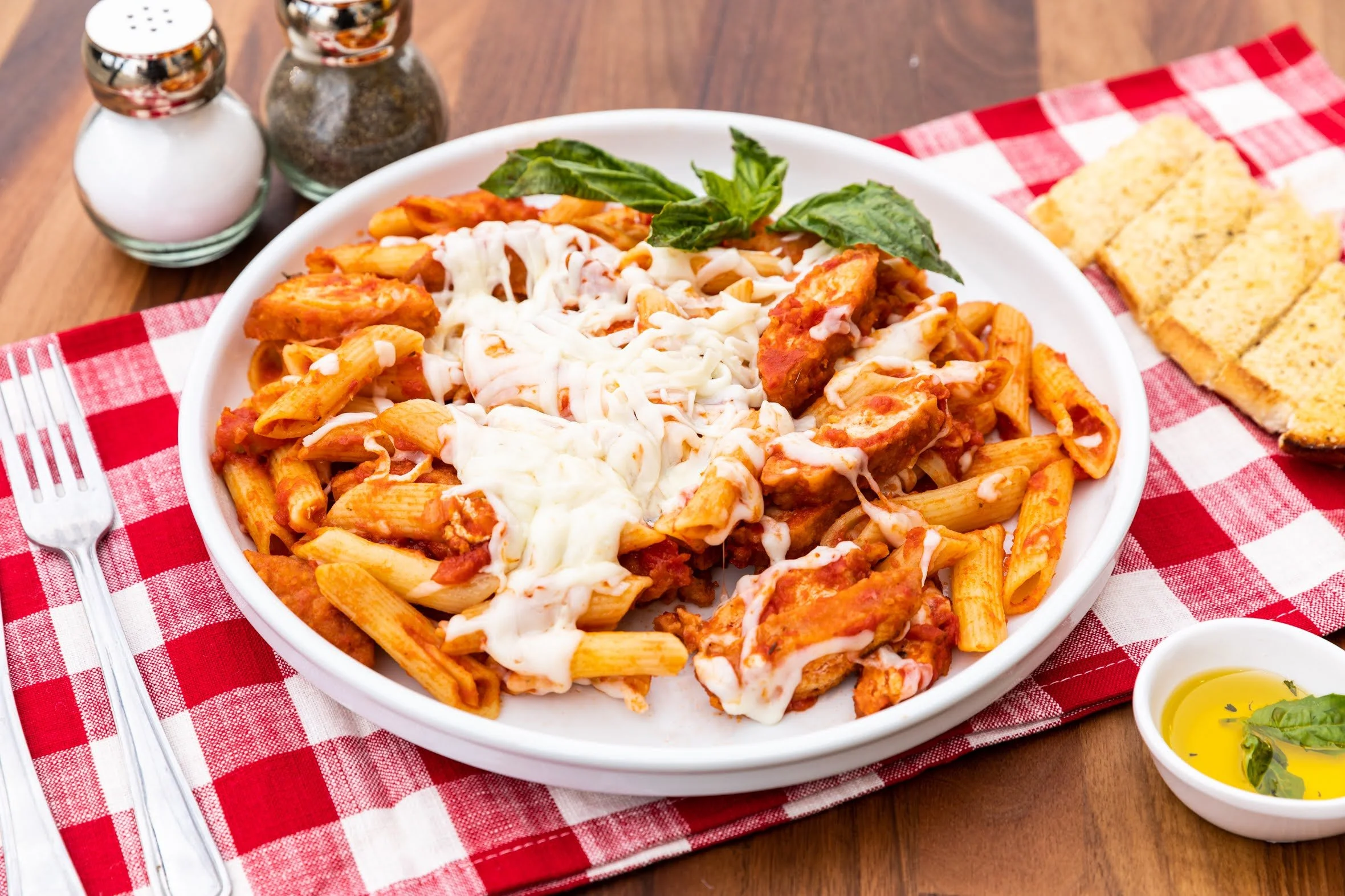 Plate of baked ziti pasta with melted cheese and basil leaves, served with garlic bread, salt, pepper, and olive oil with basil, on a red and white checkered tablecloth.