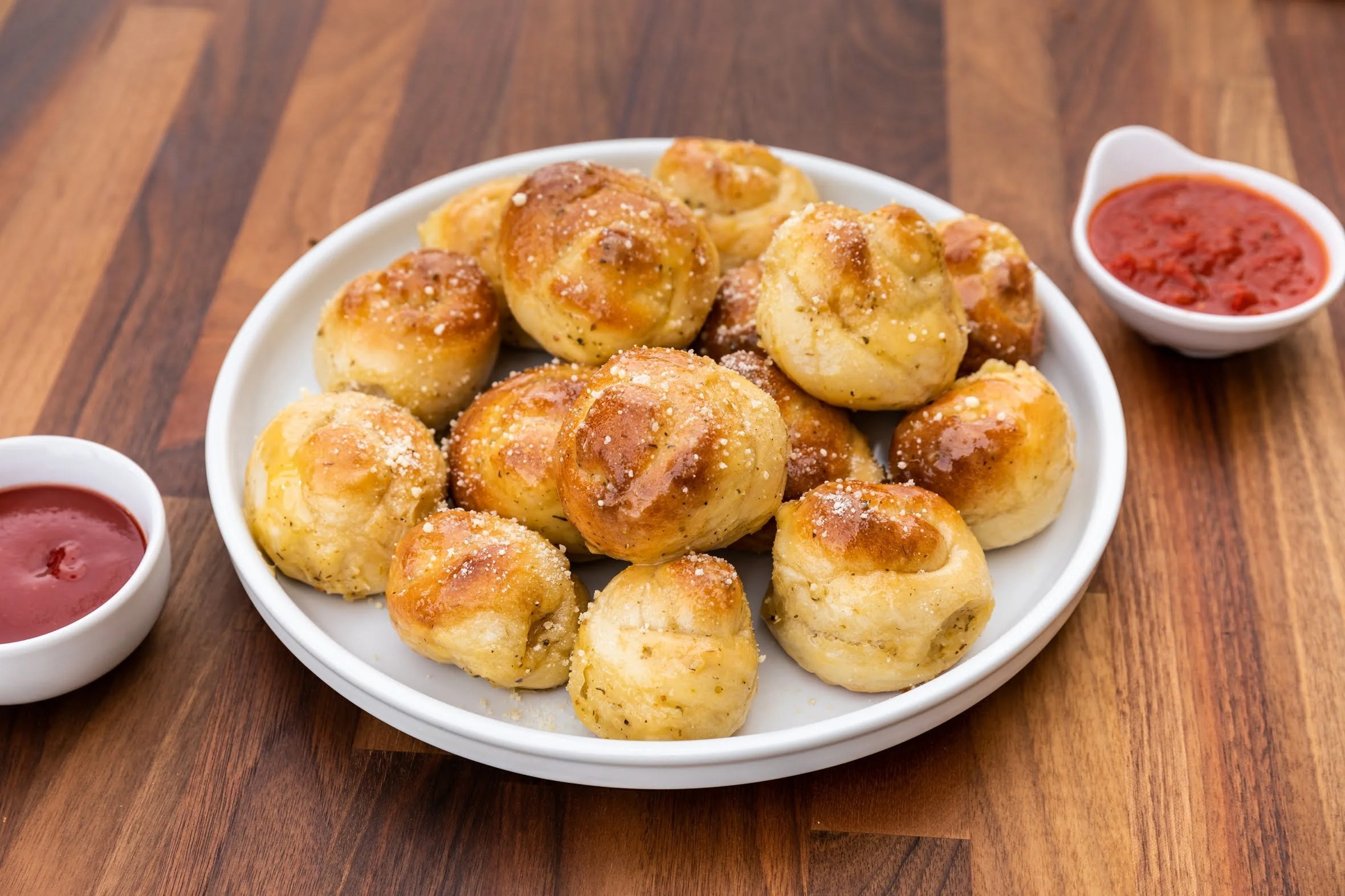 Plate of garlic bread knots sprinkled with cheese and herbs, served with two small bowls of red sauce on a wooden table.