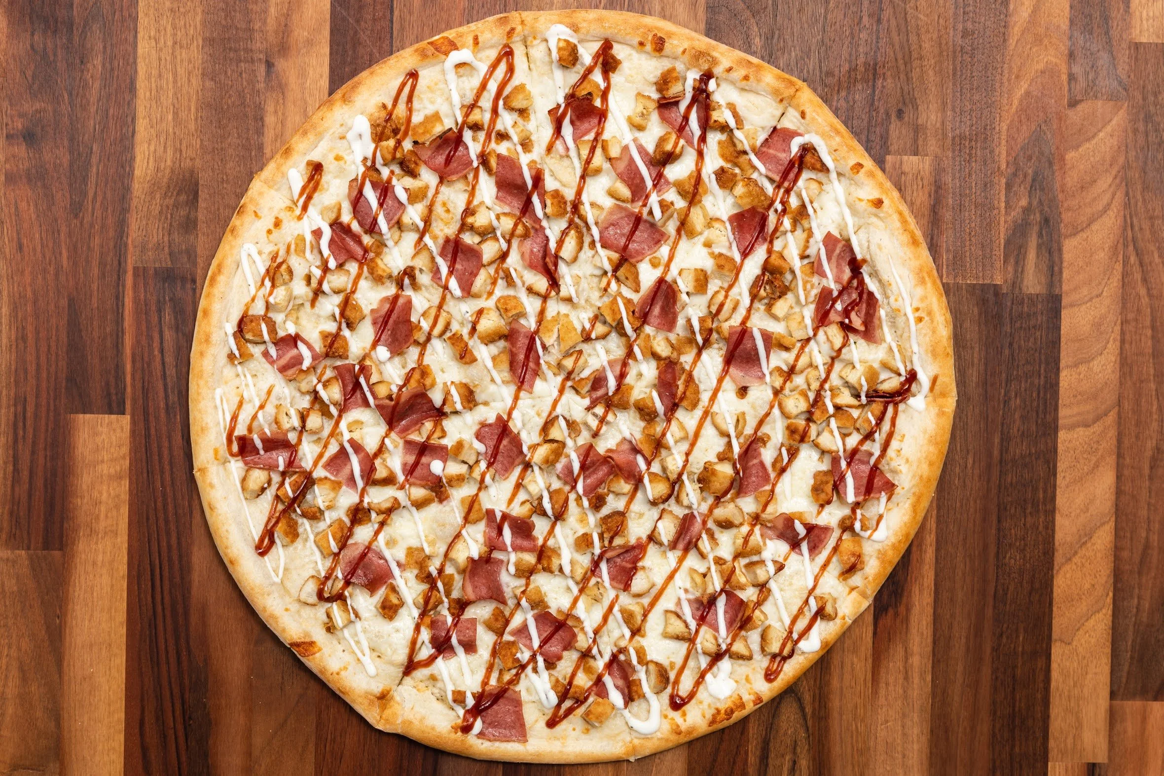 A pizza with cheese, ham, chicken, and sauces on a wooden surface.
