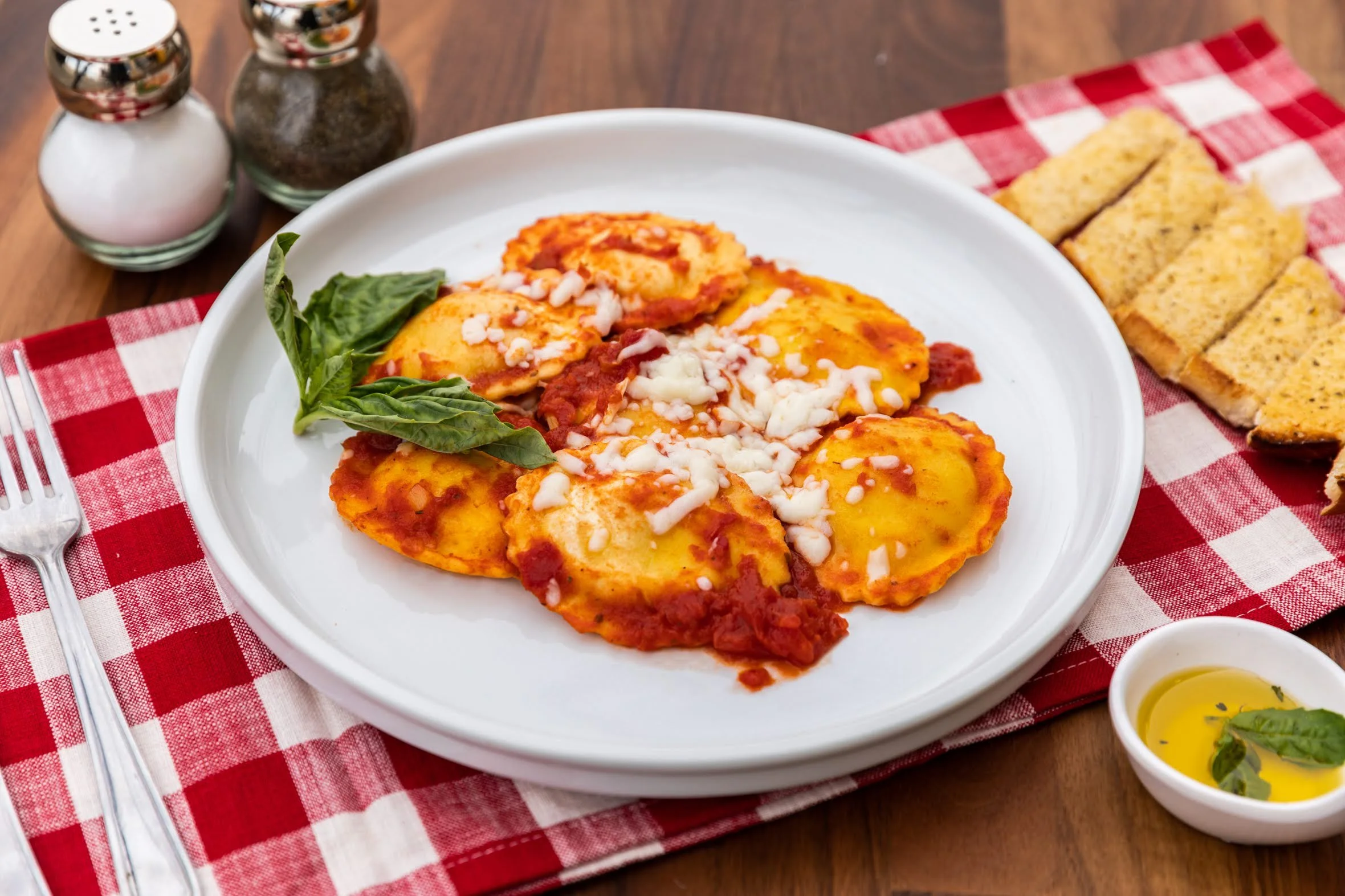 Plate of ravioli topped with tomato sauce and cheese, garnished with basil leaves, on a red and white checkered tablecloth with toasted bread, salt and pepper shakers, and a small dish of olive oil with herbs.