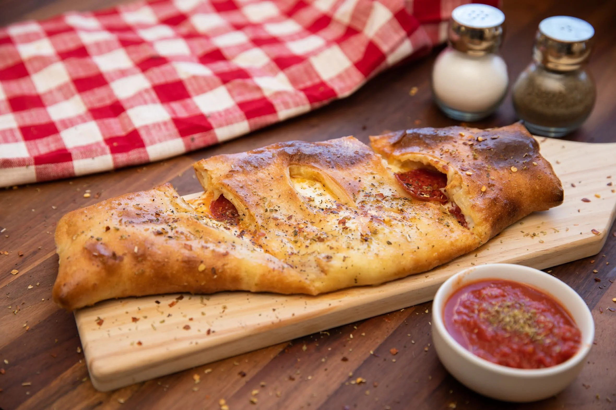 Fried calzone on a wooden serving board with a small bowl of marinara sauce, black pepper, and red pepper flakes in the background on a wooden table with a red checkered cloth