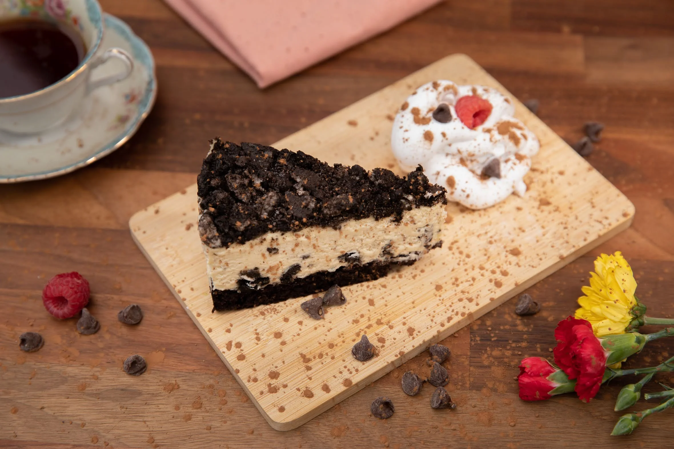 A slice of cookies and cream cheesecake with chocolate crust and crushed cookie topping, garnished with whipped cream, chocolate chips, and a raspberry on a wooden board. There is a cup of coffee, a pink napkin, and some colorful flowers nearby.